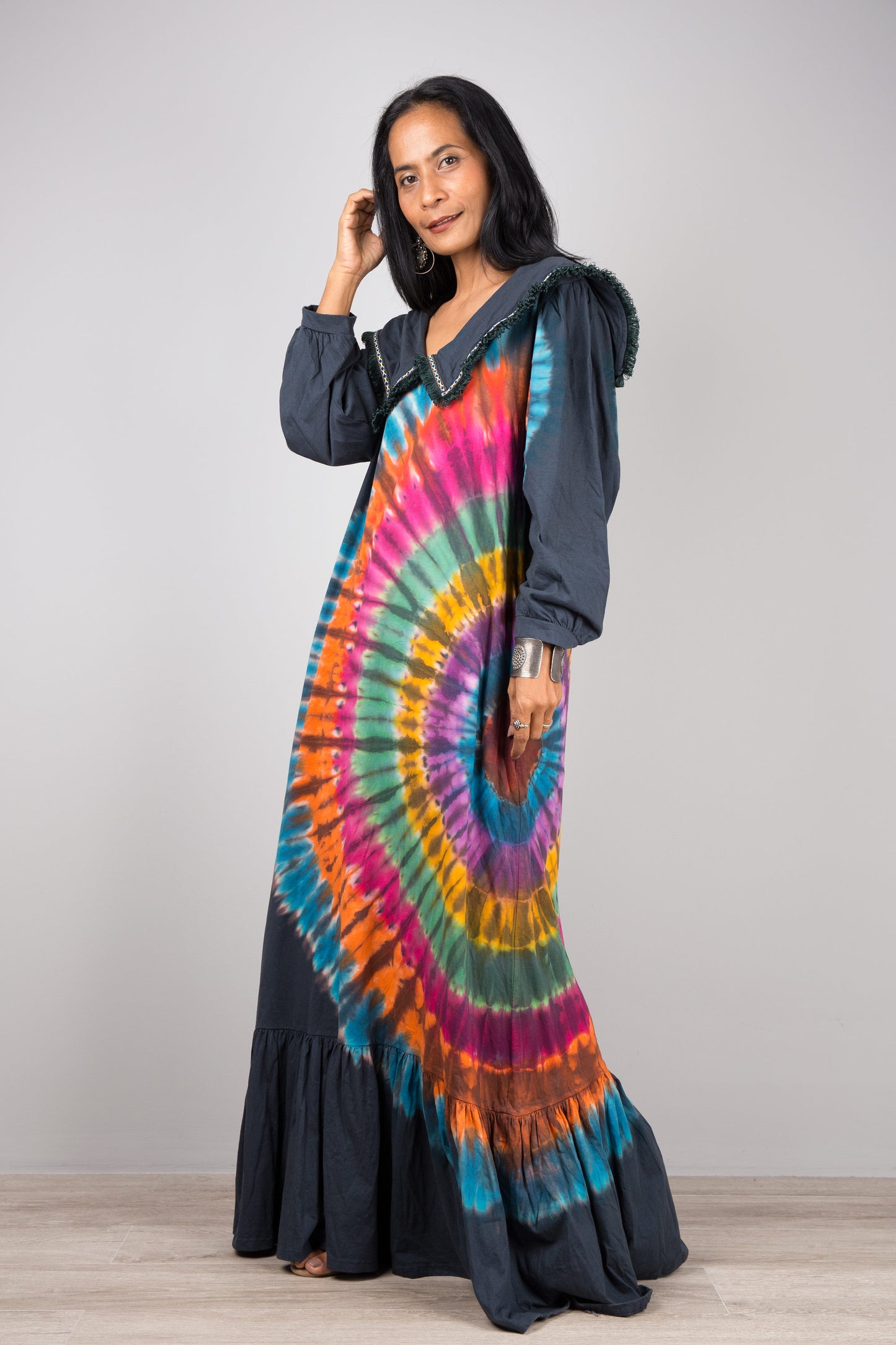 Woman showcasing a rainbow tie-dye kaftan maxi dress with navy trim, flowing design, deep V-neck, and long sleeves, accessorized with nude sandals, on a gray and wooden floor setting.