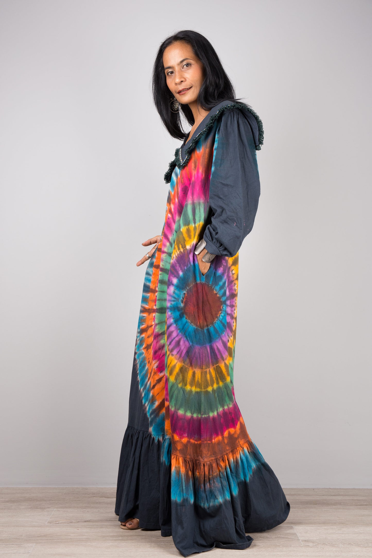 Model in a rainbow tie-dye pattern kaftan maxi dress with navy trim, deep V-neck, and long sleeves, standing on a wooden floor with a gray wall background, wearing nude sandals.