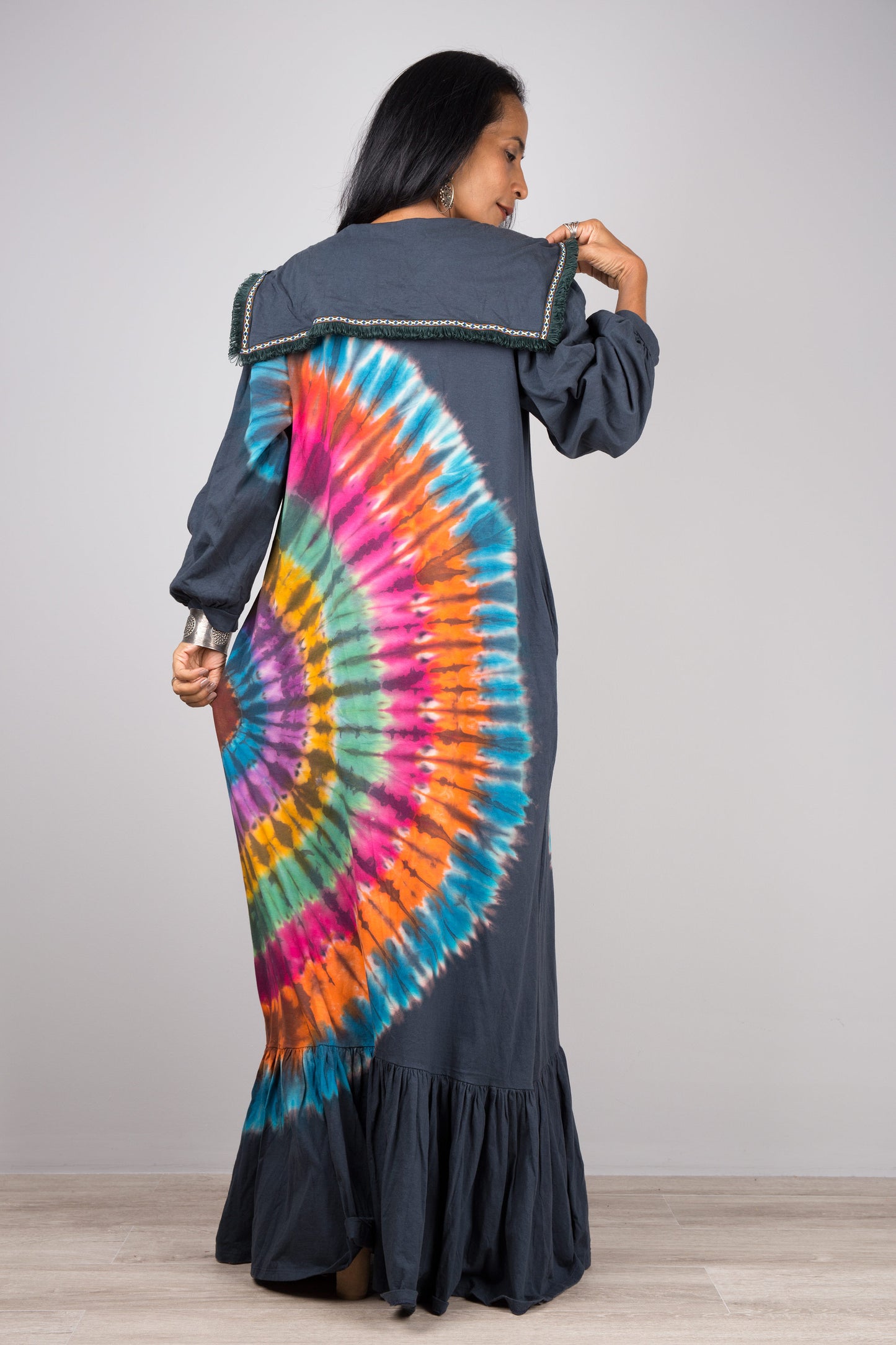 Nuichan rainbow tie-dye kaftan maxi dress with a navy-trimmed collar, deep V-neck, and long sleeves, modeled by a woman with nude sandals and dangling earrings, against a simple gray and wood backdrop.