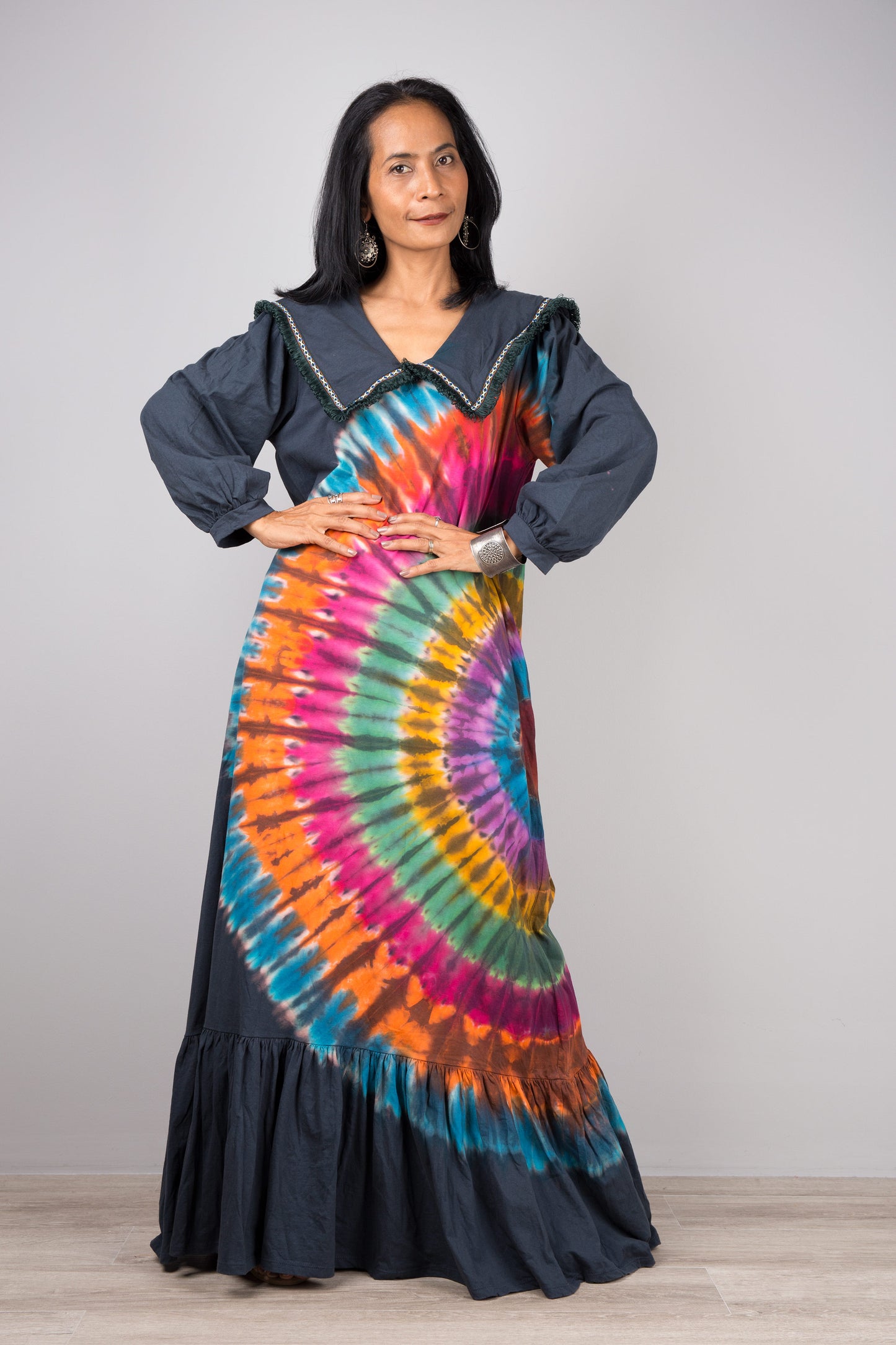 Rainbow tie-dye kaftan maxi dress with a navy-trimmed V-neck and long sleeves, worn by a woman with nude sandals and earrings, set against a minimalist gray and wood interior.