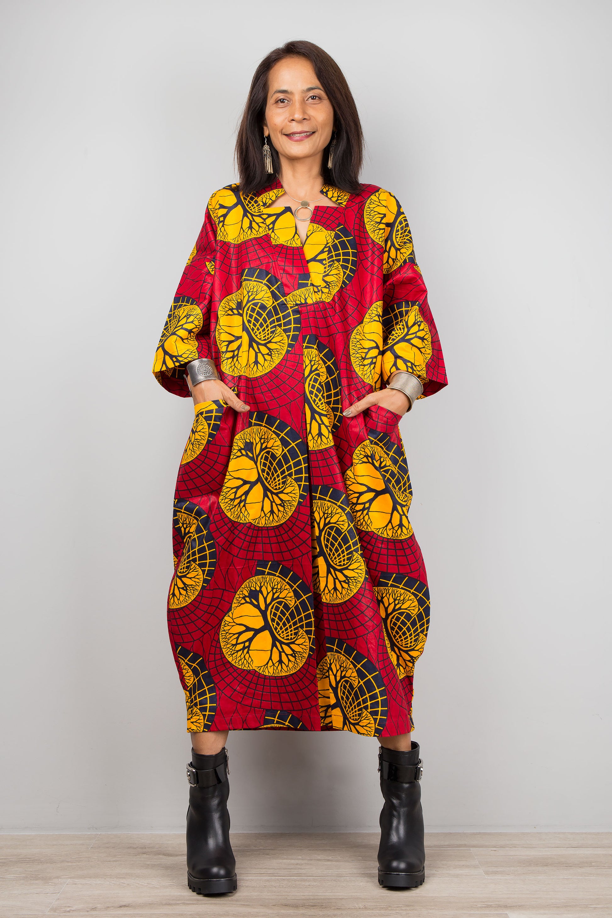 A woman wearing a red and yellow Ankara kaftan dress with a V-neck and three-quarter sleeves, standing against a gray wall with a wooden floor, accessorized with black ankle boots and silver cuffs.

