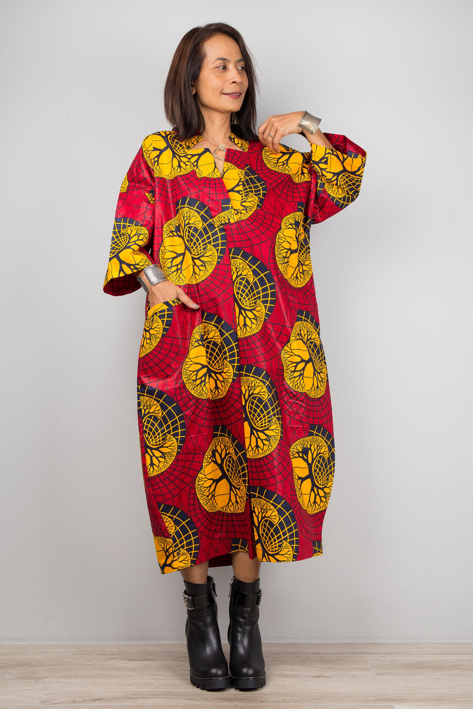 Red and yellow Ankara print kaftan dress on a woman, featuring a loose fit, V-neck, and wide sleeves, set against a minimalist gray and wood background, with black boots.


