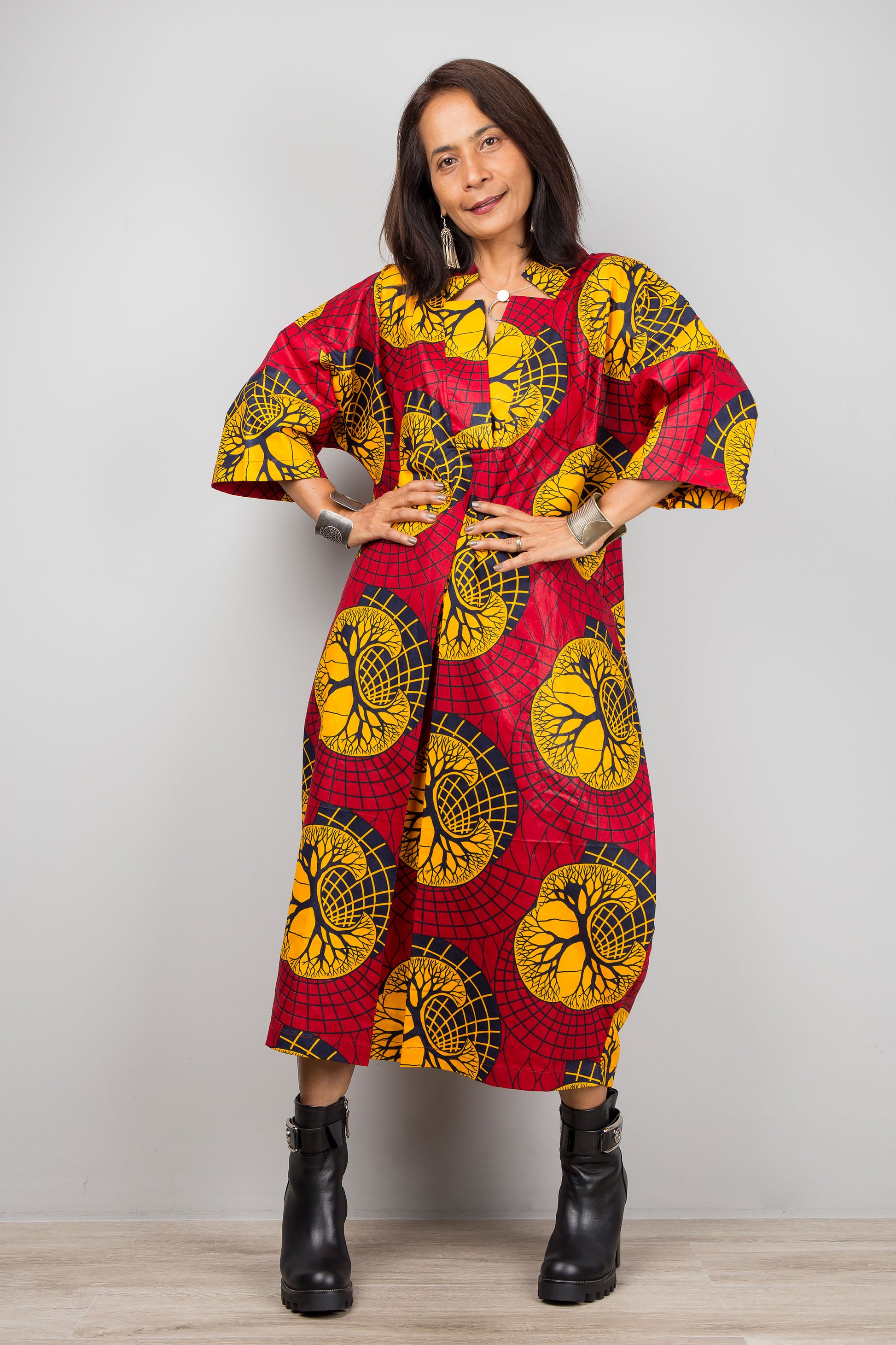 Nuichan red and yellow Ankara kaftan dress with a V-neckline and wide sleeves, modeled by a woman with silver cuffs and black boots, against a simple gray and wood backdrop.

