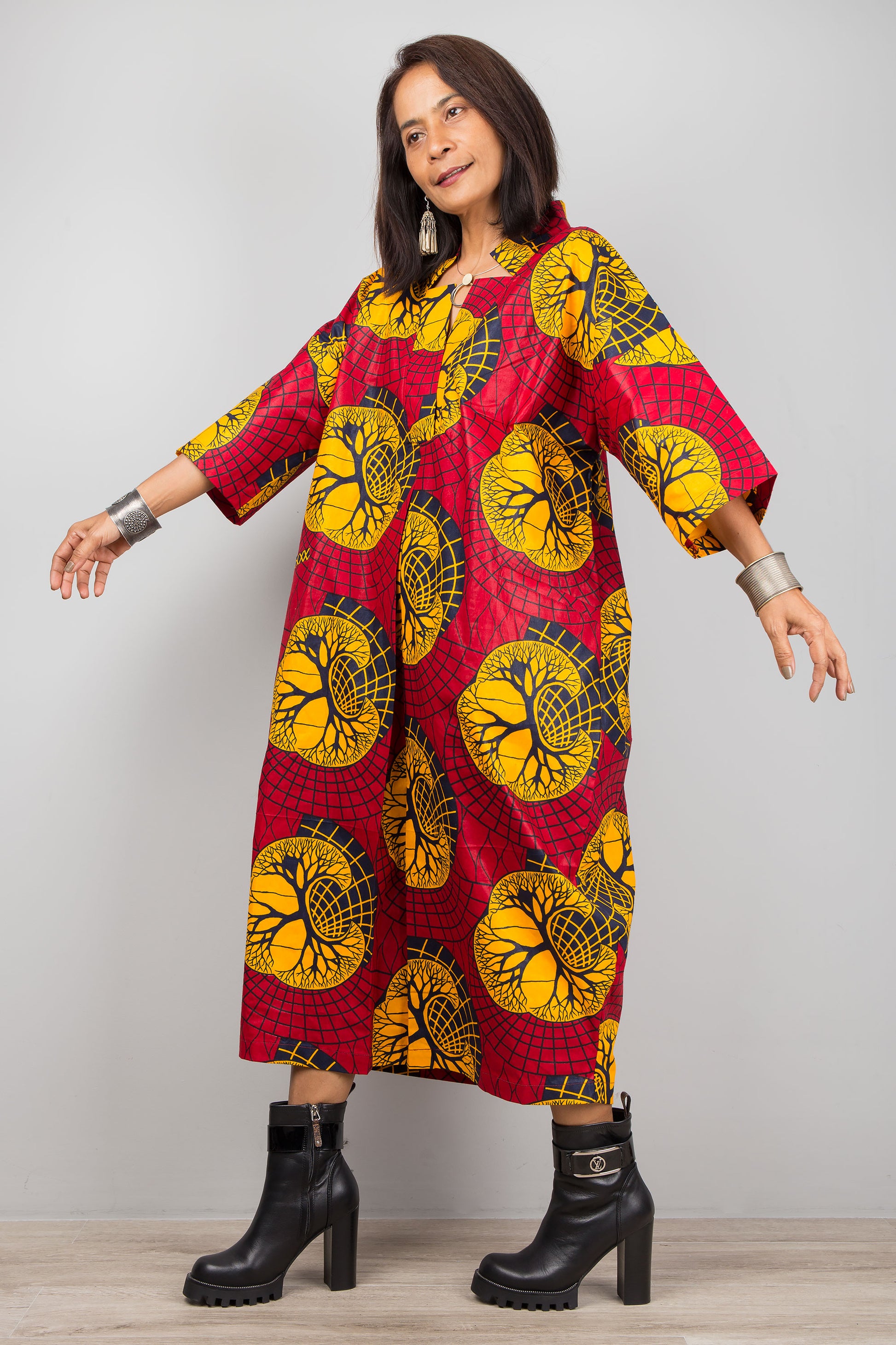 Model in a red and yellow Ankara pattern kaftan dress featuring a deep V-neck and flared three-quarter sleeves, standing on a wooden floor with a gray wall background, wearing black boots.