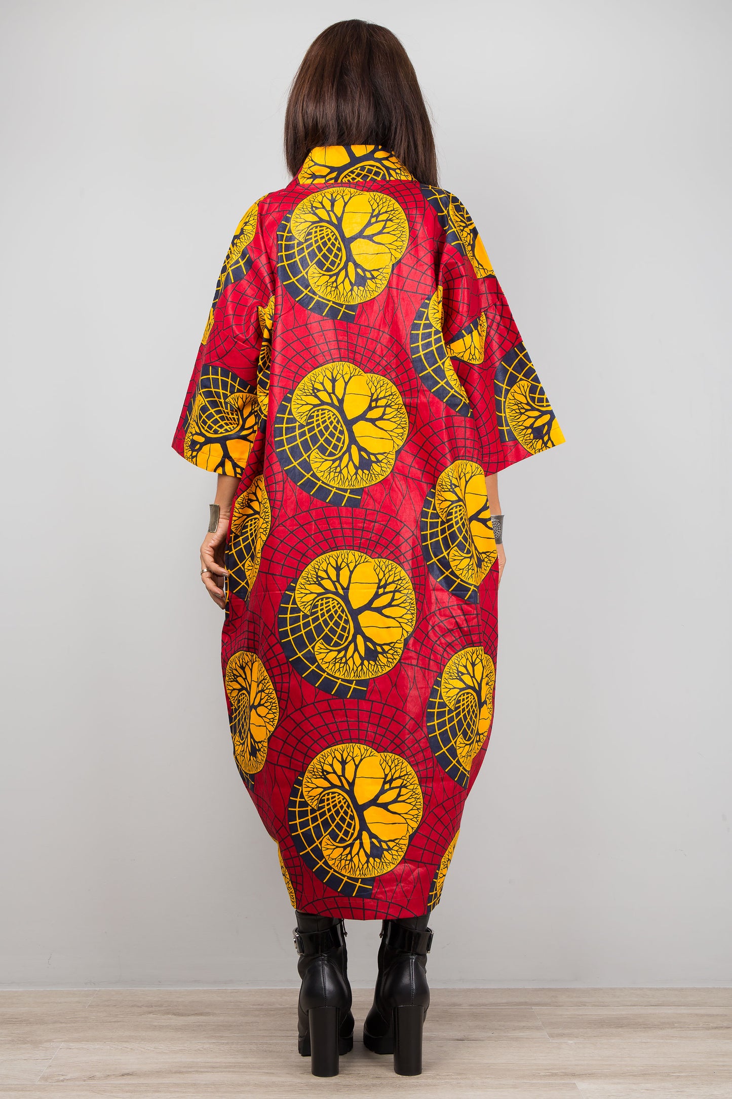 Red and yellow Ankara print kaftan dress with a deep V-neck and wide sleeves, worn by a woman with black boots and silver cuffs, set against a minimalist gray and wood interior.

