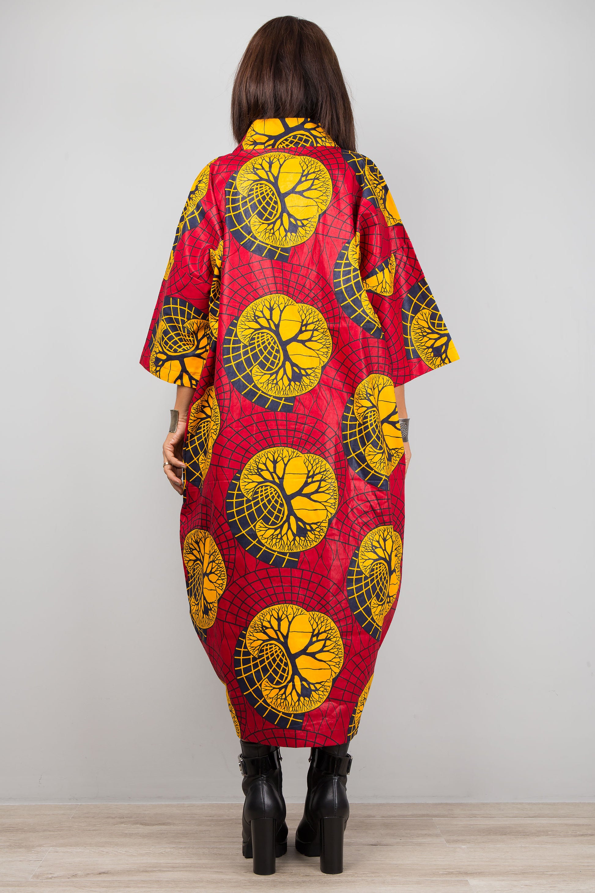 Red and yellow Ankara print kaftan dress with a deep V-neck and wide sleeves, worn by a woman with black boots and silver cuffs, set against a minimalist gray and wood interior.

