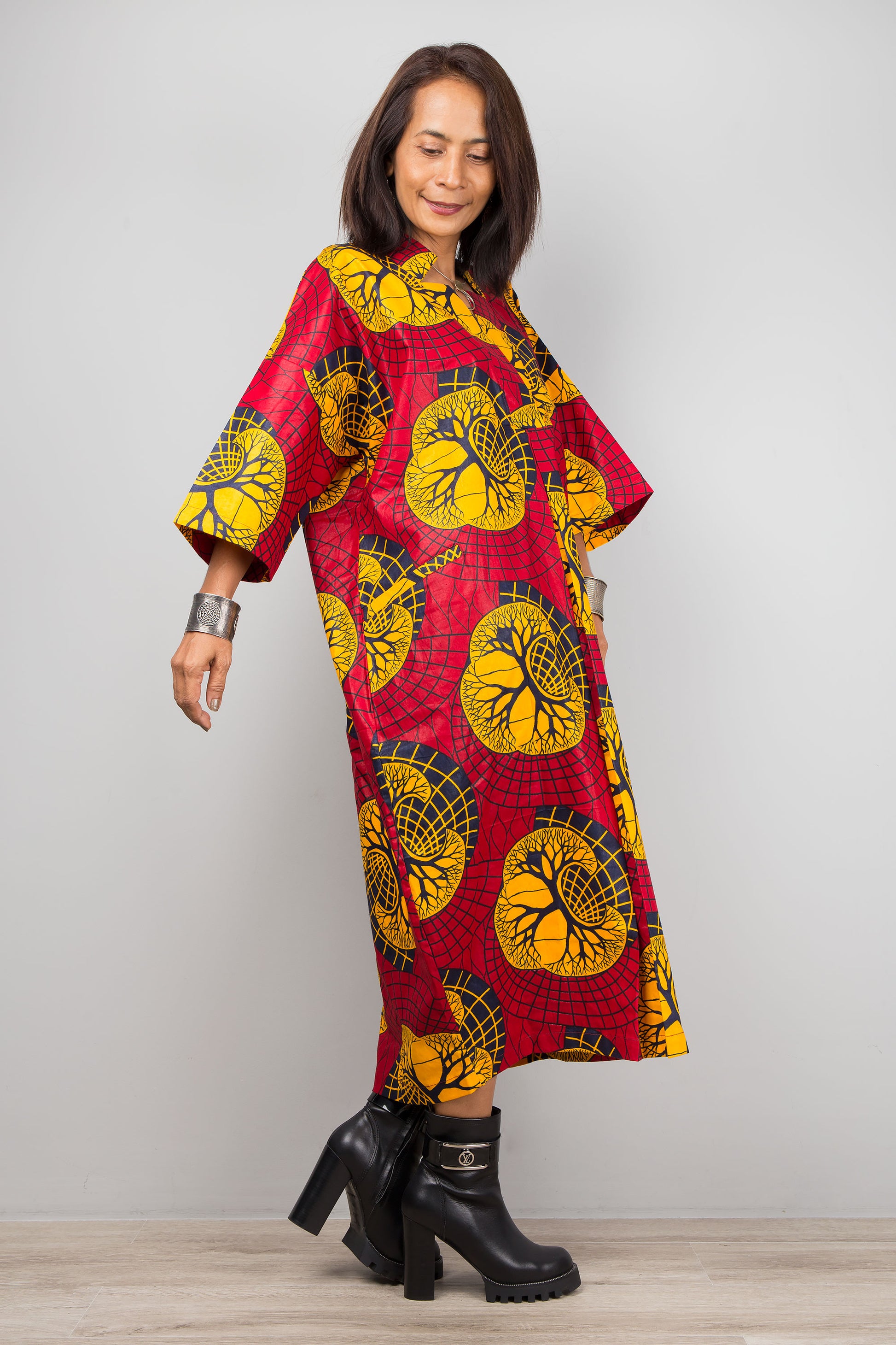 Fashionable woman wearing a red and yellow Ankara kaftan dress with a V-neck and three-quarter sleeves, standing on a wooden floor with a gray wall backdrop, accessorized with black boots.


