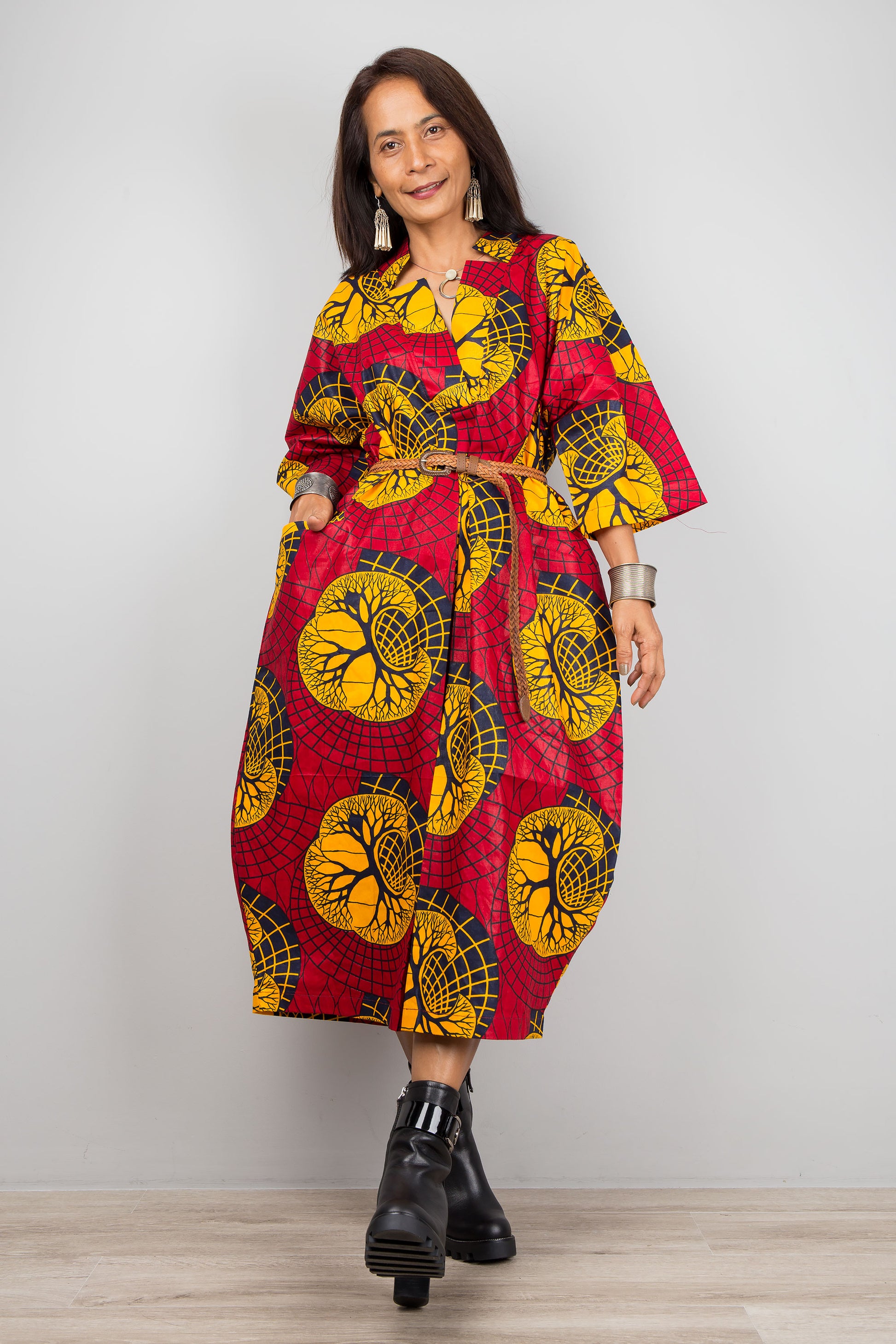 Woman showcasing a red and yellow Ankara print kaftan dress with a flowing design, V-neck, and three-quarter sleeves, accessorized with black boots and silver jewelry, on a gray and wooden floor.

