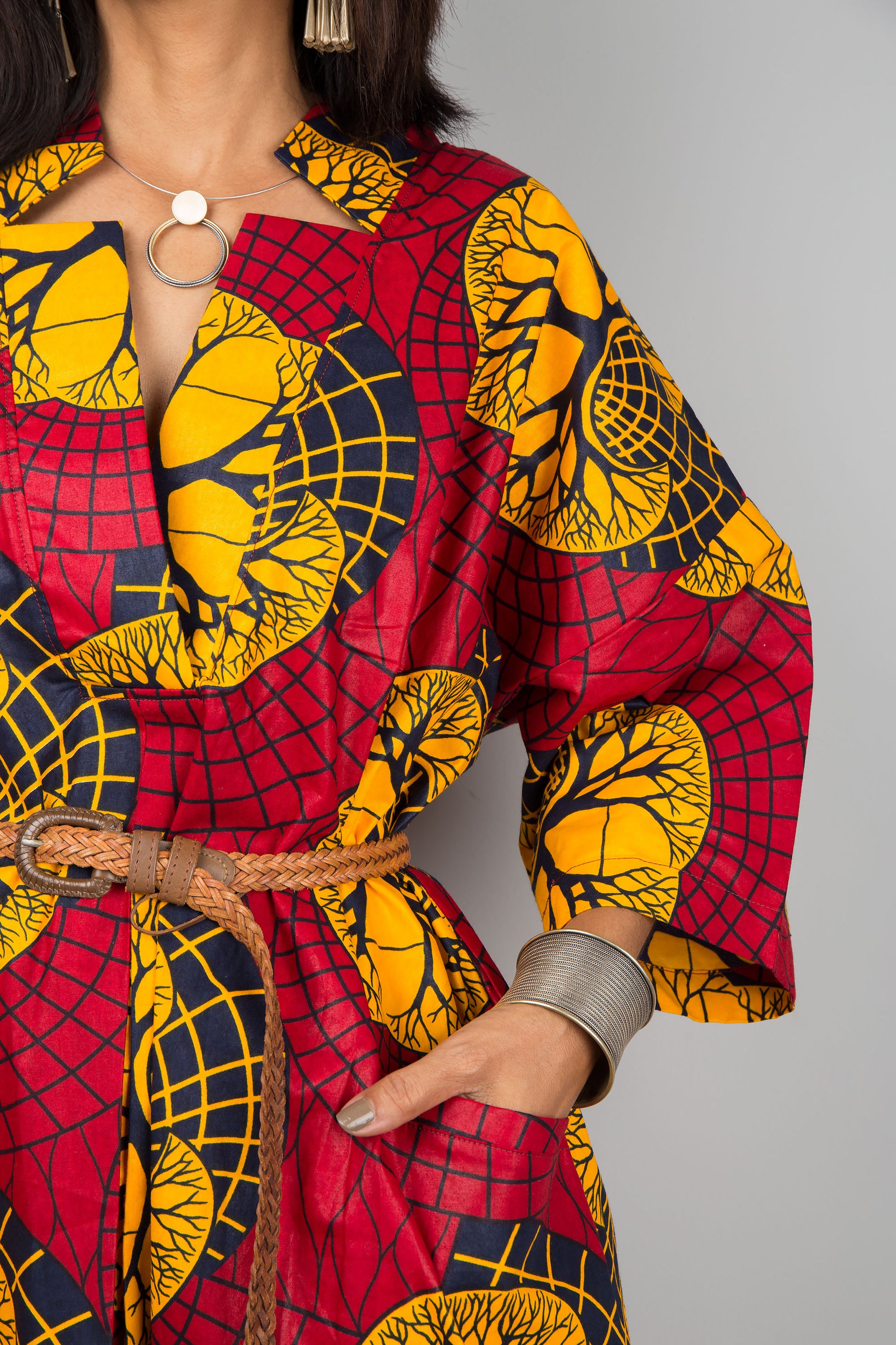 Elegant red and yellow Ankara kaftan dress with a V-neck and wide sleeves, worn by a woman against a gray wall and wooden floor, paired with black ankle boots and silver cuffs.

