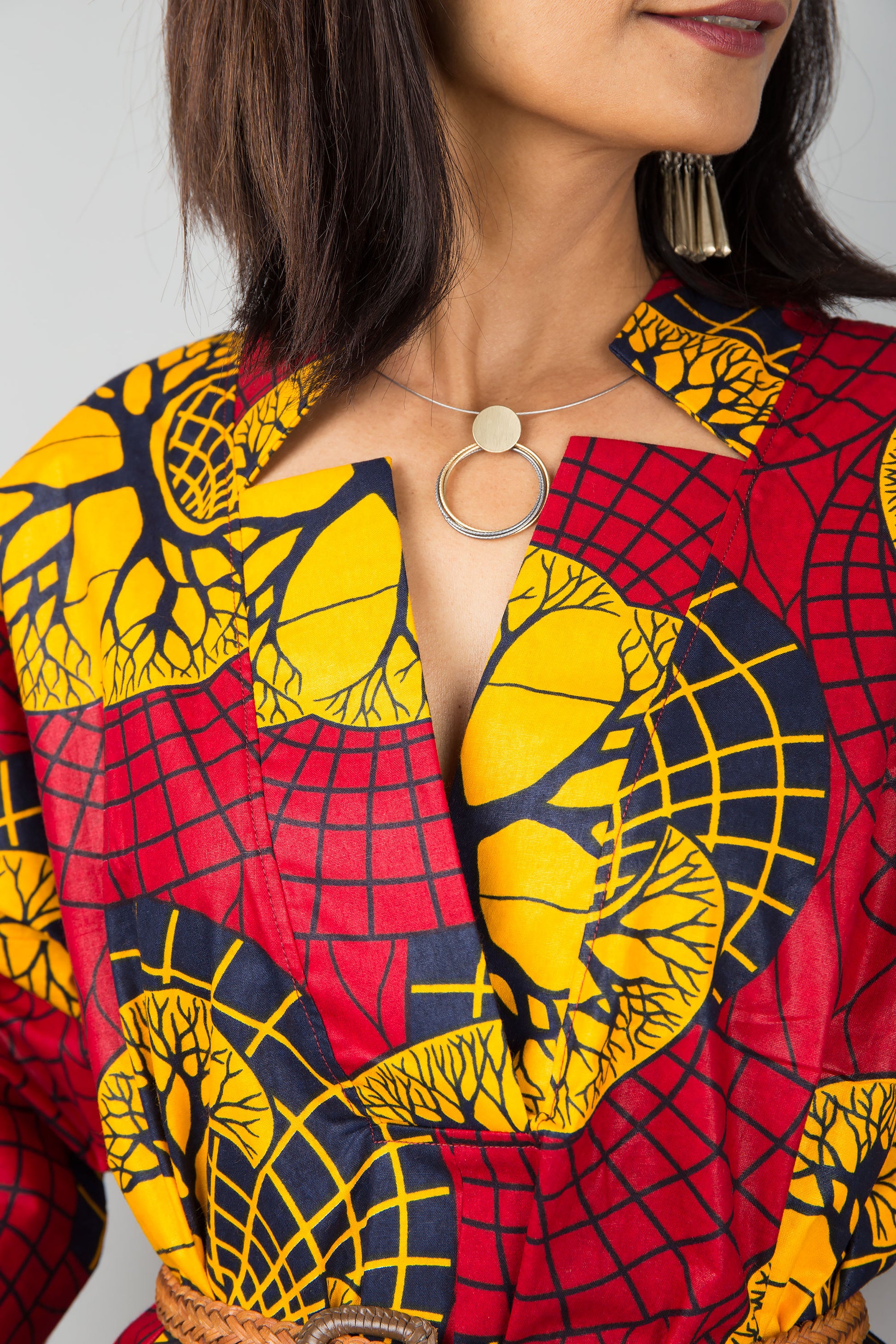 Stylish woman in a midi-length red and yellow Ankara kaftan dress with a V-neck and flared sleeves, posed against a gray wall and wooden floor, wearing black ankle boots.

