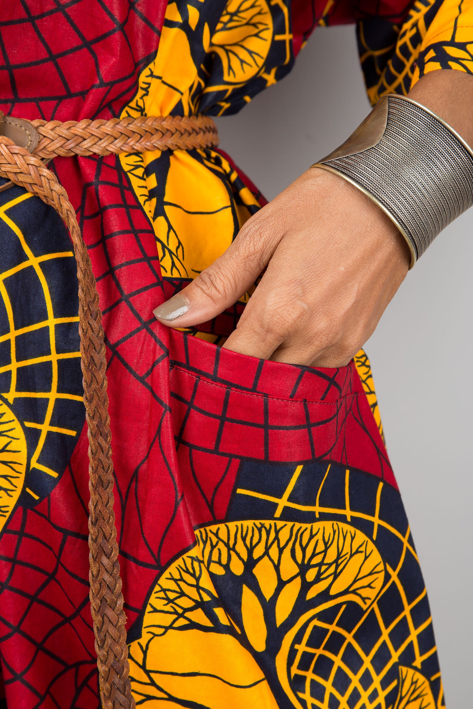 Woman in a vibrant red and yellow Ankara kaftan dress with a V-neck and loose sleeves, posed against a gray wall and wooden floor, styled with black ankle boots and silver accessories.

