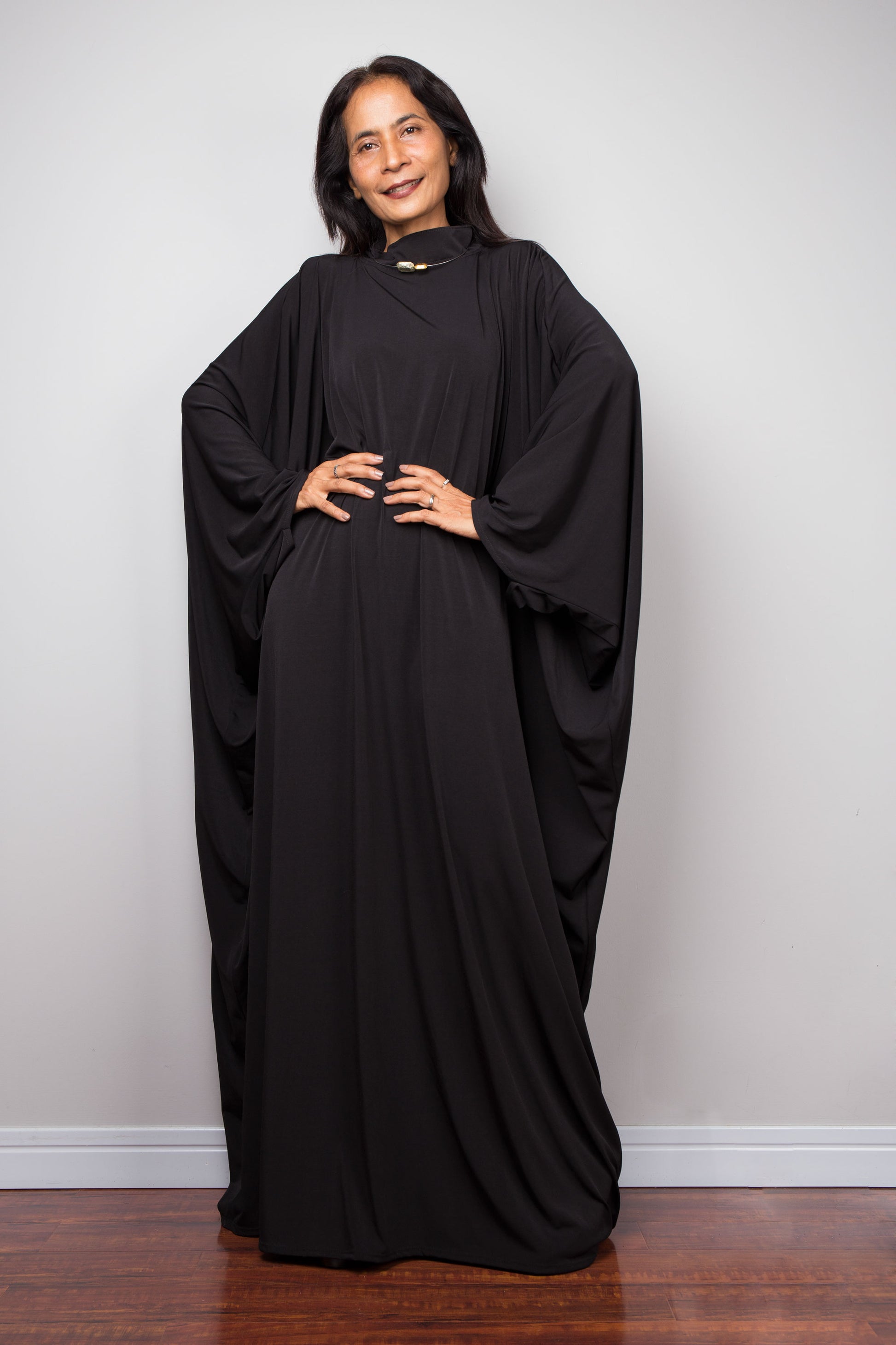 Woman wearing a black abaya standing against a plain white wall. Black kaftan dress by Nuichan