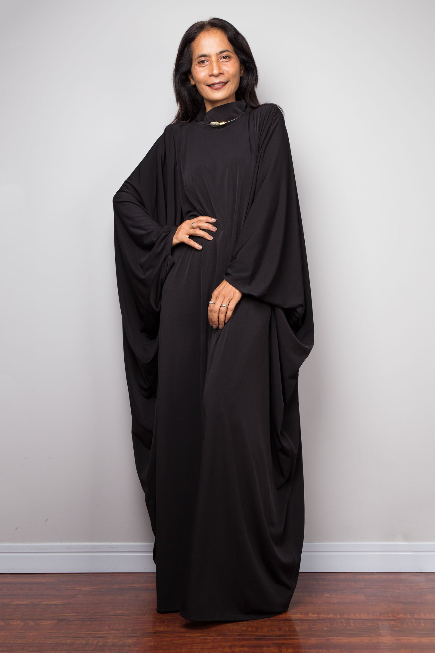 Woman wearing a black abaya against a plain white wall. Plus size black caftan by Nuichan