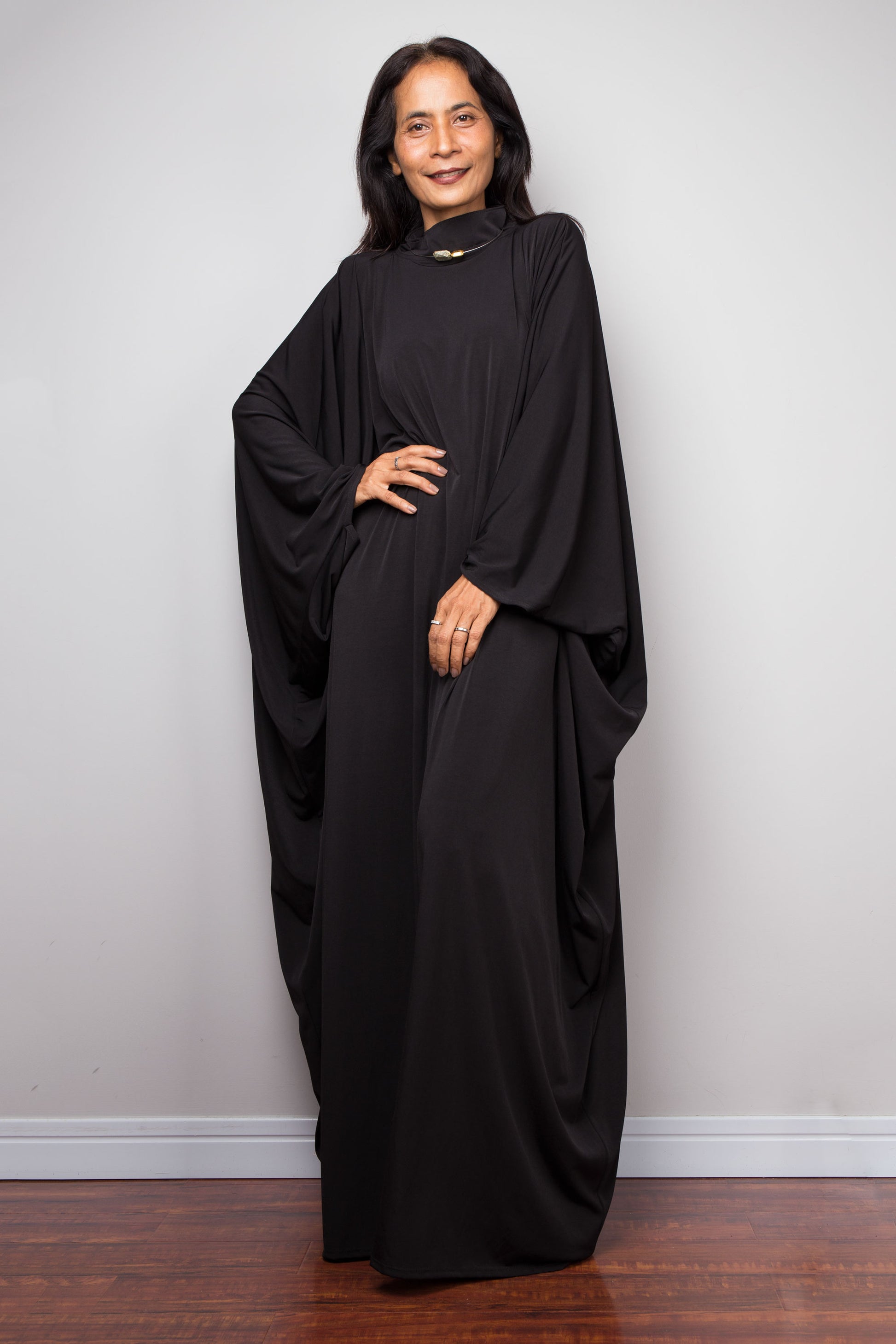 Woman wearing a black abaya against a plain white wall. Plus size black caftan by Nuichan