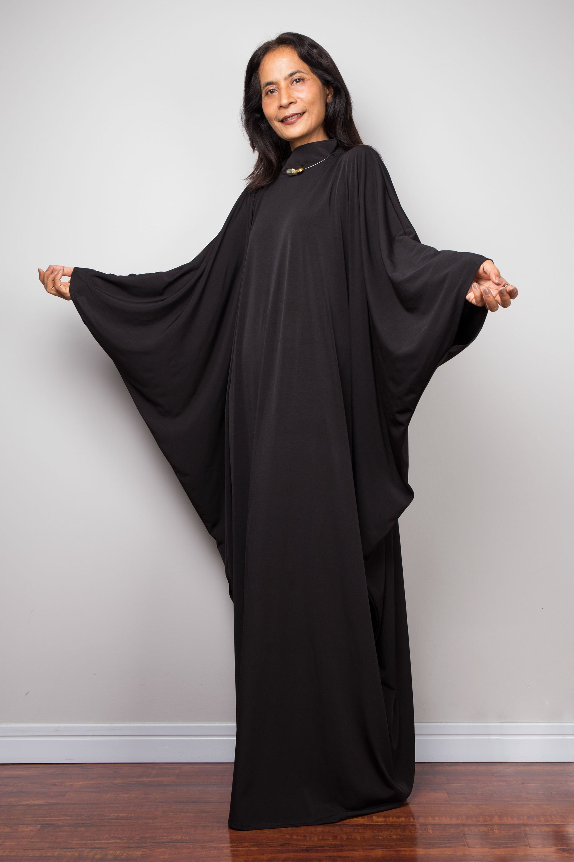 Woman wearing a black abaya standing against a plain white wall. Black kaftan dress by Nuichan