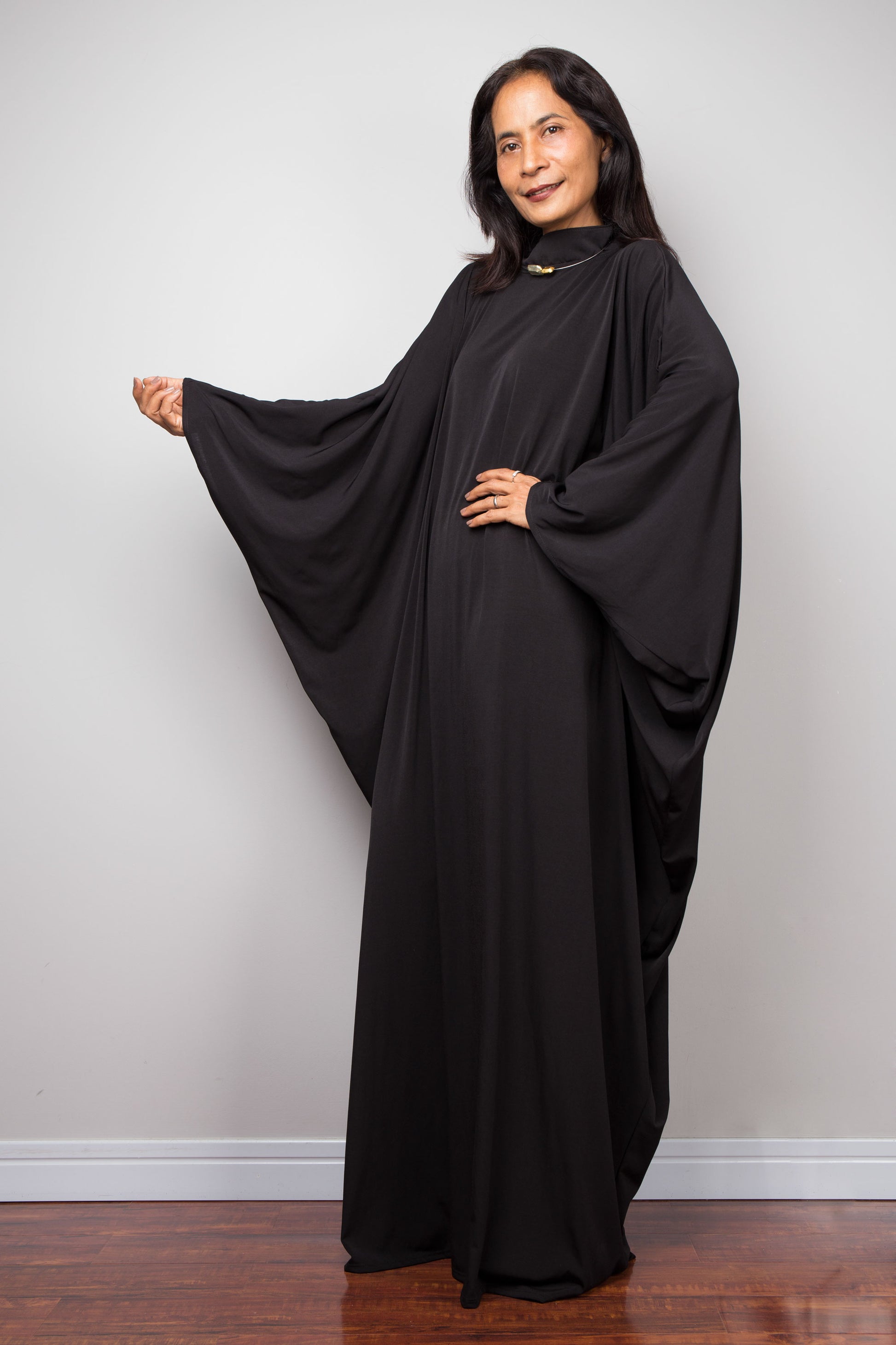 Woman wearing a black abaya standing against a plain wall. Plus size black kaftan by Nuichan