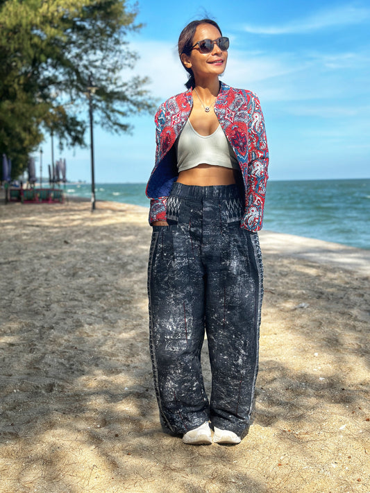 A person standing on a beach wearing black tie-dye batik hemp pants with a pattern that includes floral and geometric designs, paired with a multicolored top and sunglasses.