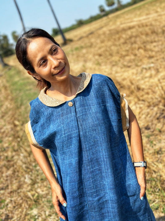 Nuichan’s blue hemp kaftan dress with brown earth-tone batik patterns, modeled by a woman with a ponytail, posed outdoors in a golden field with palm trees, wearing a smartwatch and featuring a Peter Pan collar.

