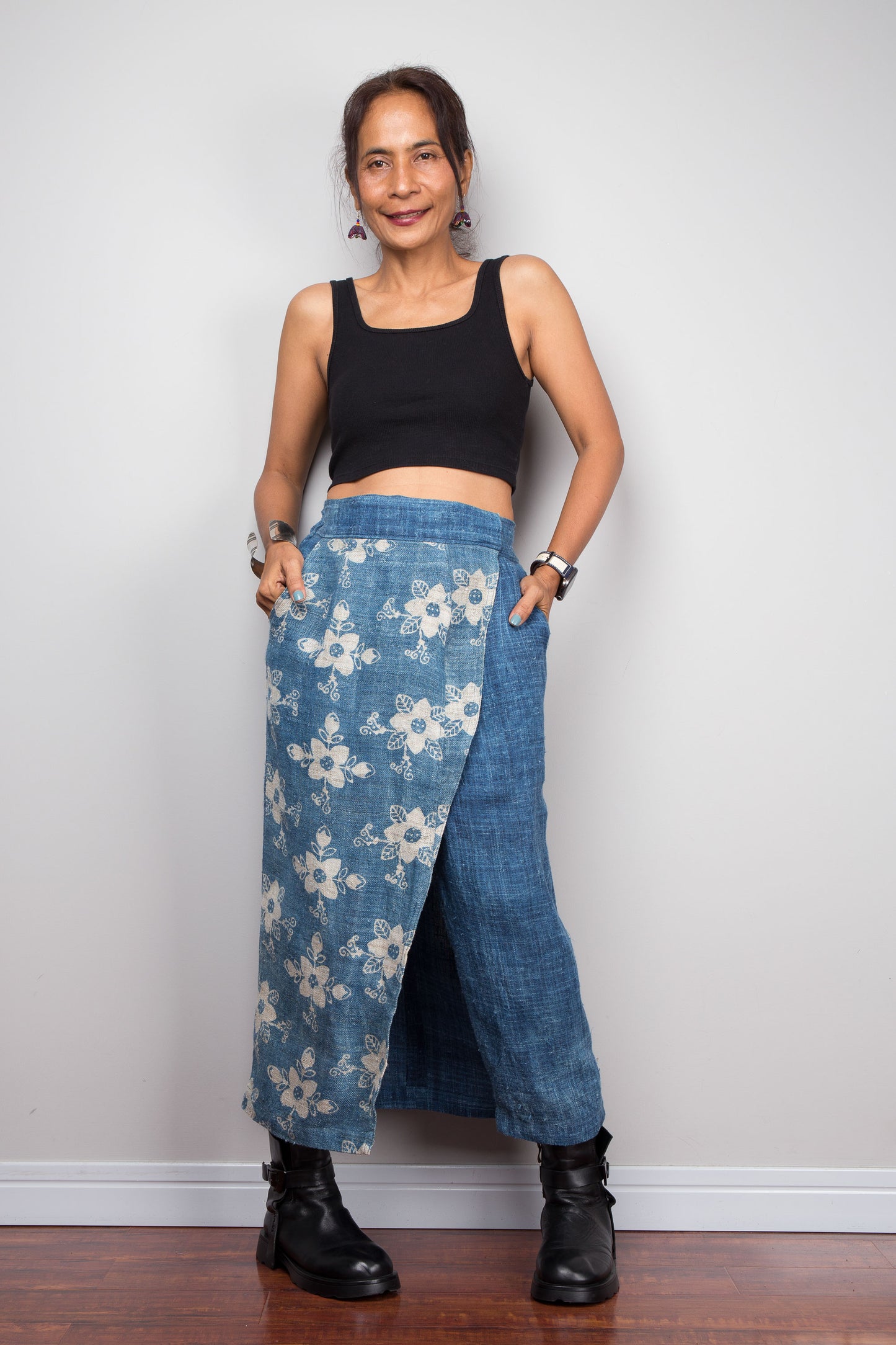 Front-facing view of a petite female model wearing the Nuichan indigo blue handwoven hemp wrap skirt with hands in side pockets, showcasing the hand block printed floral pattern on the front wrap panel in natural cream on indigo blue, wrap front construction with front split opening, and elastic waistband, styled with a black crop top and ankle boots