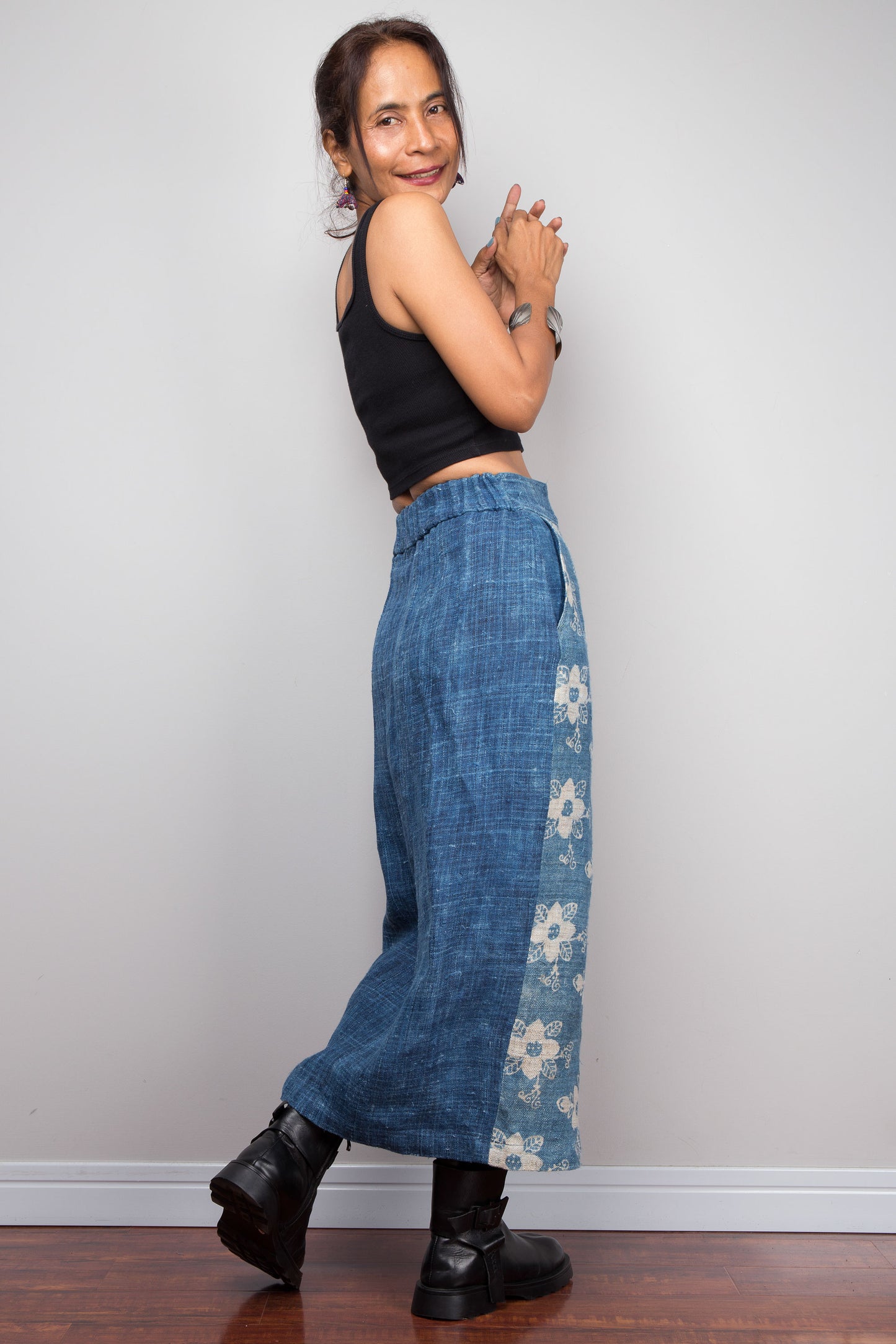 Three quarter back-facing view of a model wearing the Nuichan indigo blue handwoven hemp wrap skirt showcasing the hand block printed floral pattern on the front wrap panel in natural cream on indigo blue, wrap front construction with front split opening, and elastic waistband, styled with a black crop top and ankle boots