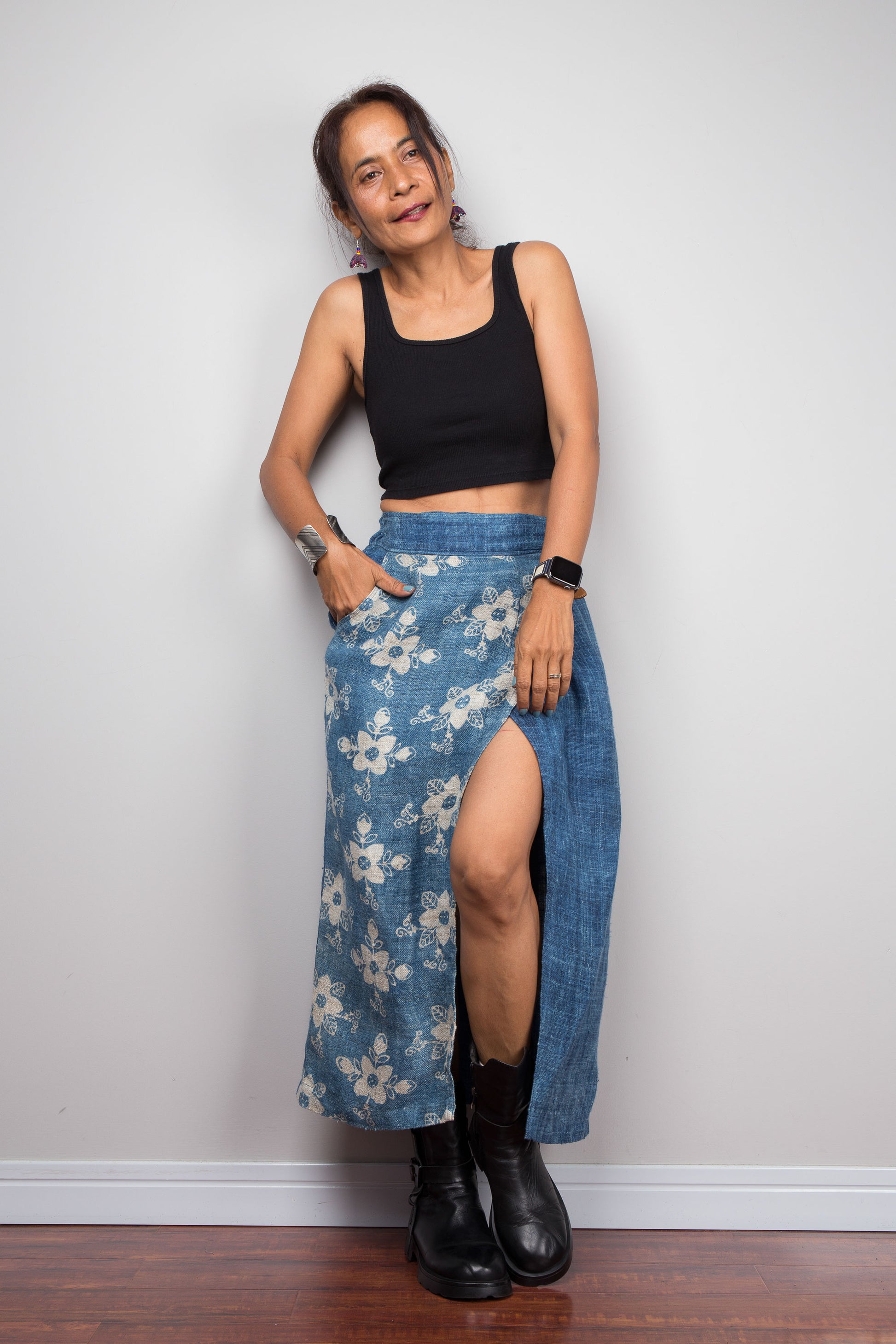 Front view of a petite female model wearing the Nuichan indigo blue handwoven hemp wrap skirt with hands in side pockets, showcasing the hand block printed floral pattern on the front wrap panel in natural cream on indigo blue, wrap front construction with front split opening, and elastic waistband, styled with a black crop top and ankle boots
