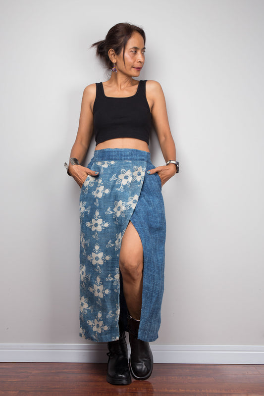 Front-facing view of a petite model wearing the Nuichan indigo blue handwoven hemp wrap skirt with hands in side pockets, showcasing the hand block printed floral pattern on the front wrap panel in natural cream on indigo blue, wrap front construction with front split opening, and elastic waistband, styled with a black crop top and ankle boots