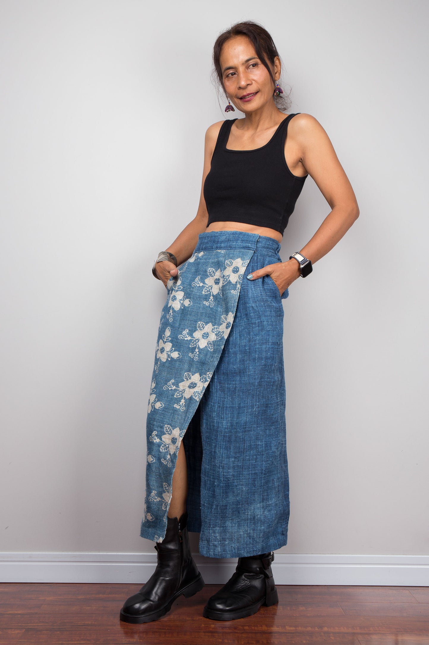 Three quarter view of a model wearing the Nuichan indigo blue handwoven hemp wrap skirt with hands in side pockets, showcasing the hand block printed floral pattern on the front wrap panel in natural cream on indigo blue, wrap front construction with front split opening, and elastic waistband, styled with a black crop top and ankle boots
