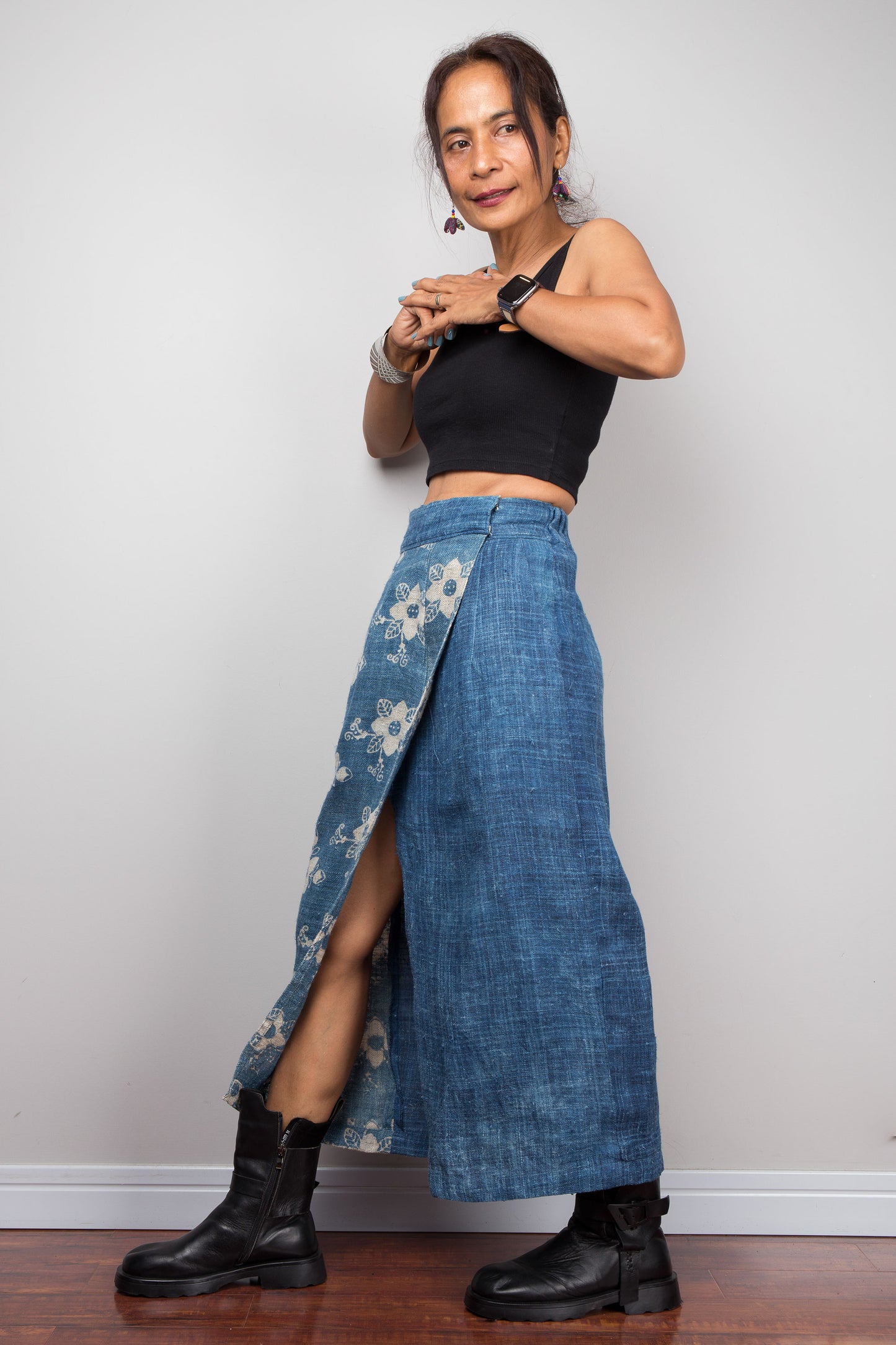 Side facing view of a model wearing the Nuichan indigo blue handwoven hemp petite wrap skirt, showcasing the hand block printed floral pattern on the front wrap panel in natural cream on indigo blue, wrap front construction with front split opening, and elastic waistband, styled with a black crop top and ankle boots