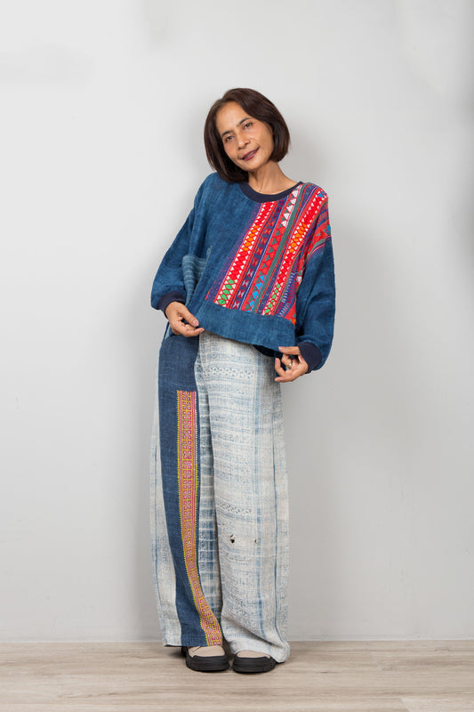 Woman wearing a blue hemp sweater with colorful patterns and white pants with multicolor stripes against a plain background