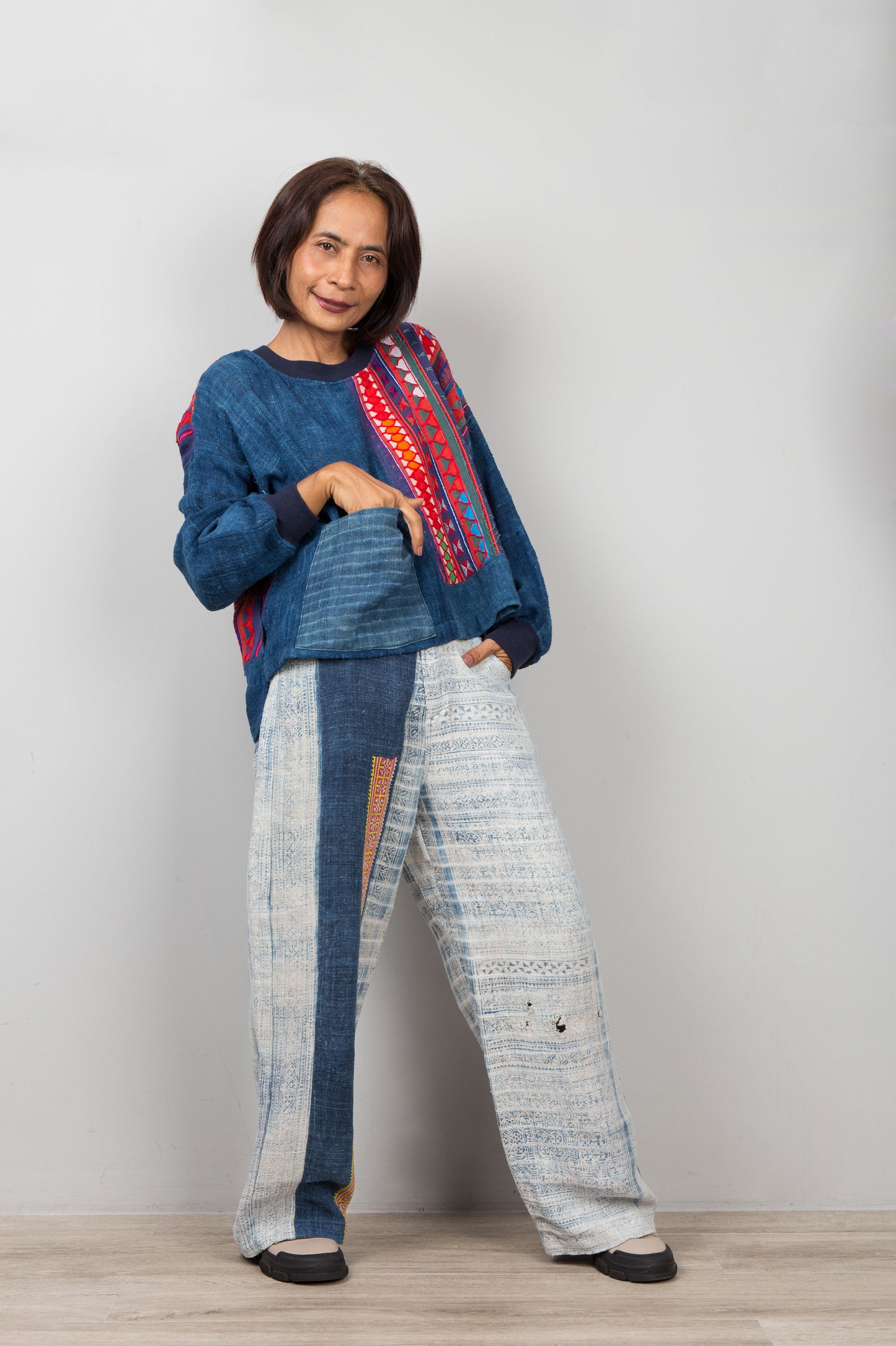 Person wearing a blue hemp patchwork sweater with colorful patterns and white pants against a plain background