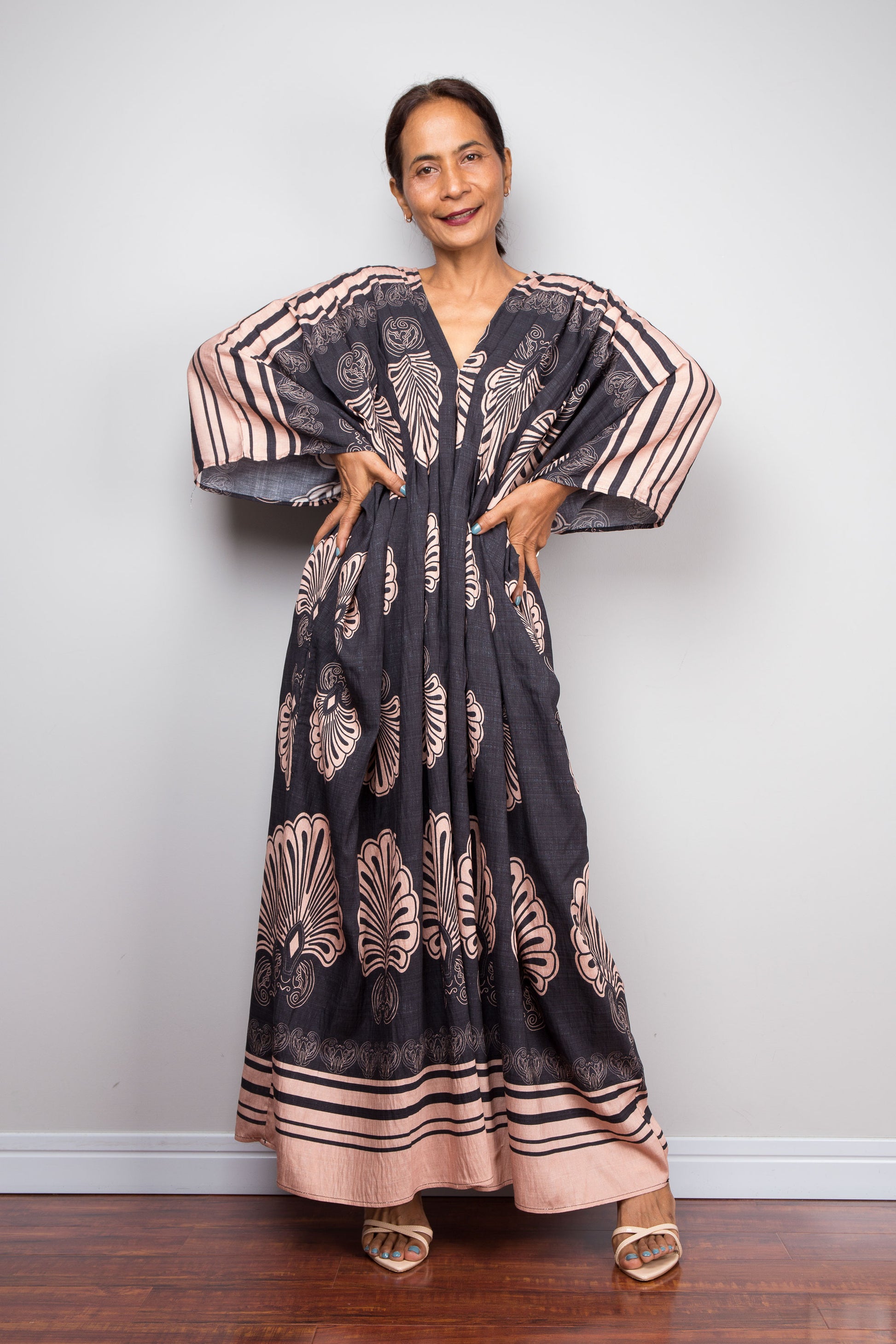 Front-facing view of a petite model wearing the Nuichan charcoal and blush Art Deco batwing kaftan dress with hands on hips, showcasing the dramatic shoulder pleating, wide kimono sleeves with striped border, and bold fan print