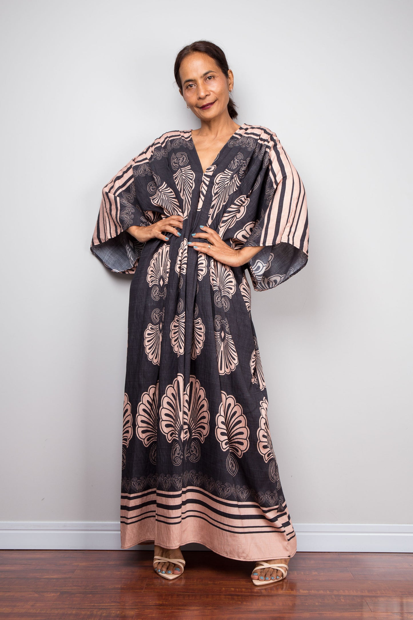 Front-facing studio shot of a petite female model in the Nuichan charcoal and blush batwing kaftan dress with hands on hips, displaying the wide kimono sleeves with contrasting underside print, deep V-neckline, and dramatic shoulder pleating