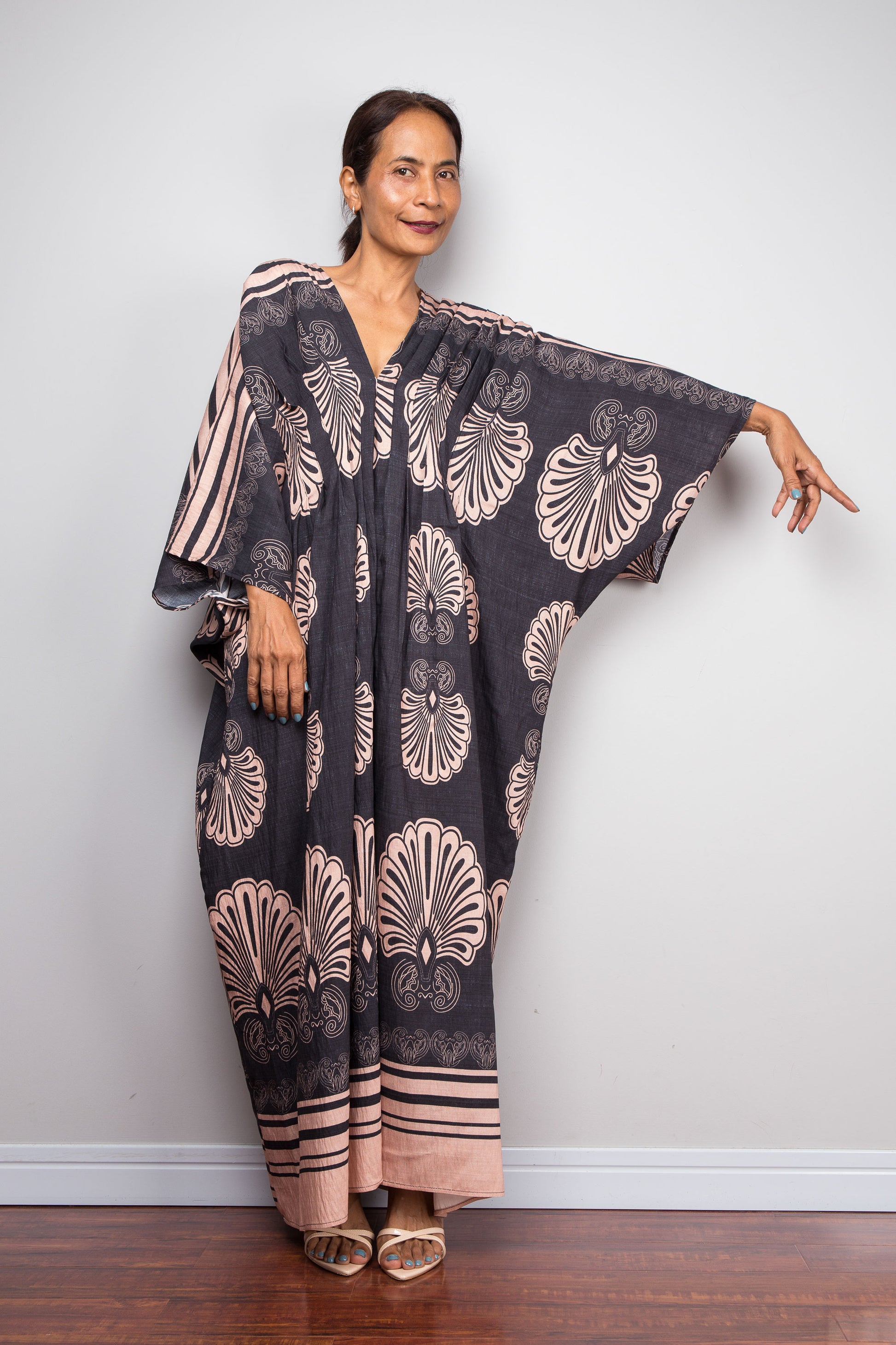 Front-facing view of a petite model wearing the Nuichan charcoal and blush Art Deco batwing kaftan dress with arm extended, showcasing the dramatic shoulder pleating, wide kimono sleeves with striped border, and bold fan print