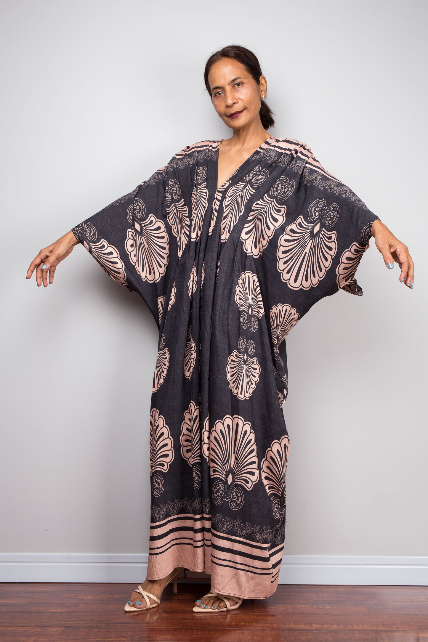 Front-facing view of a model wearing the Nuichan charcoal and blush Art Deco batwing kaftan dress with arm extended, showcasing the dramatic shoulder pleating, wide kimono sleeves with striped border, and bold fan print