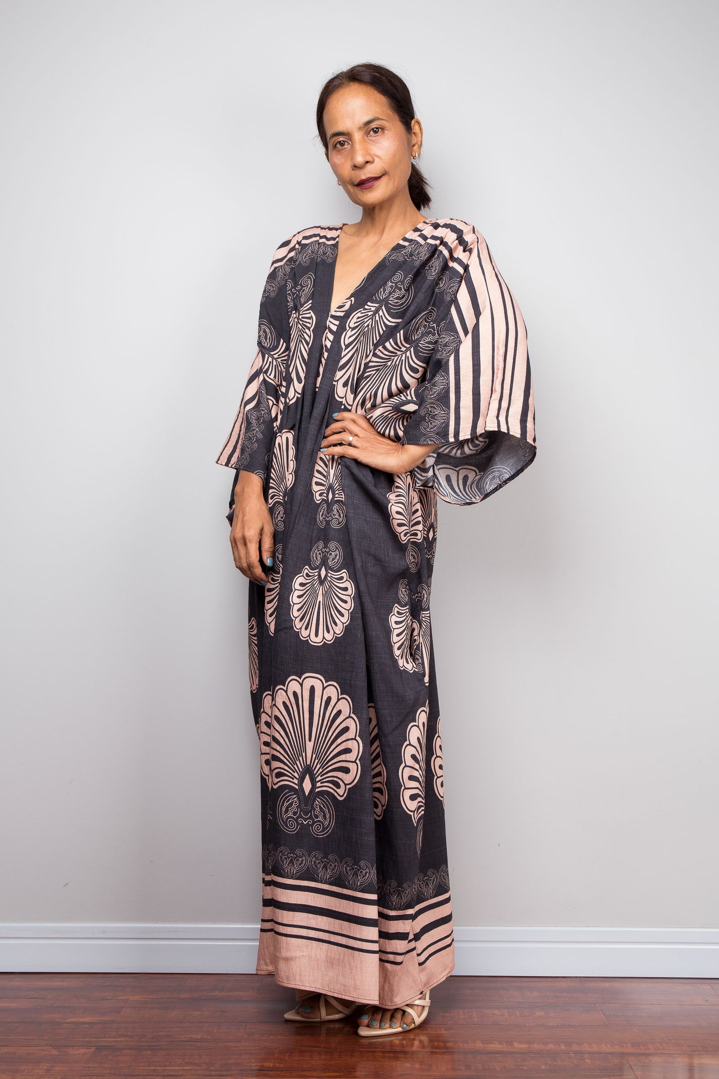 Three quarter shot of a petite model in the Nuichan charcoal and blush batwing kaftan dress with hand on hip, displaying the wide kimono sleeves with contrasting underside print, deep V-neckline, and dramatic shoulder pleating