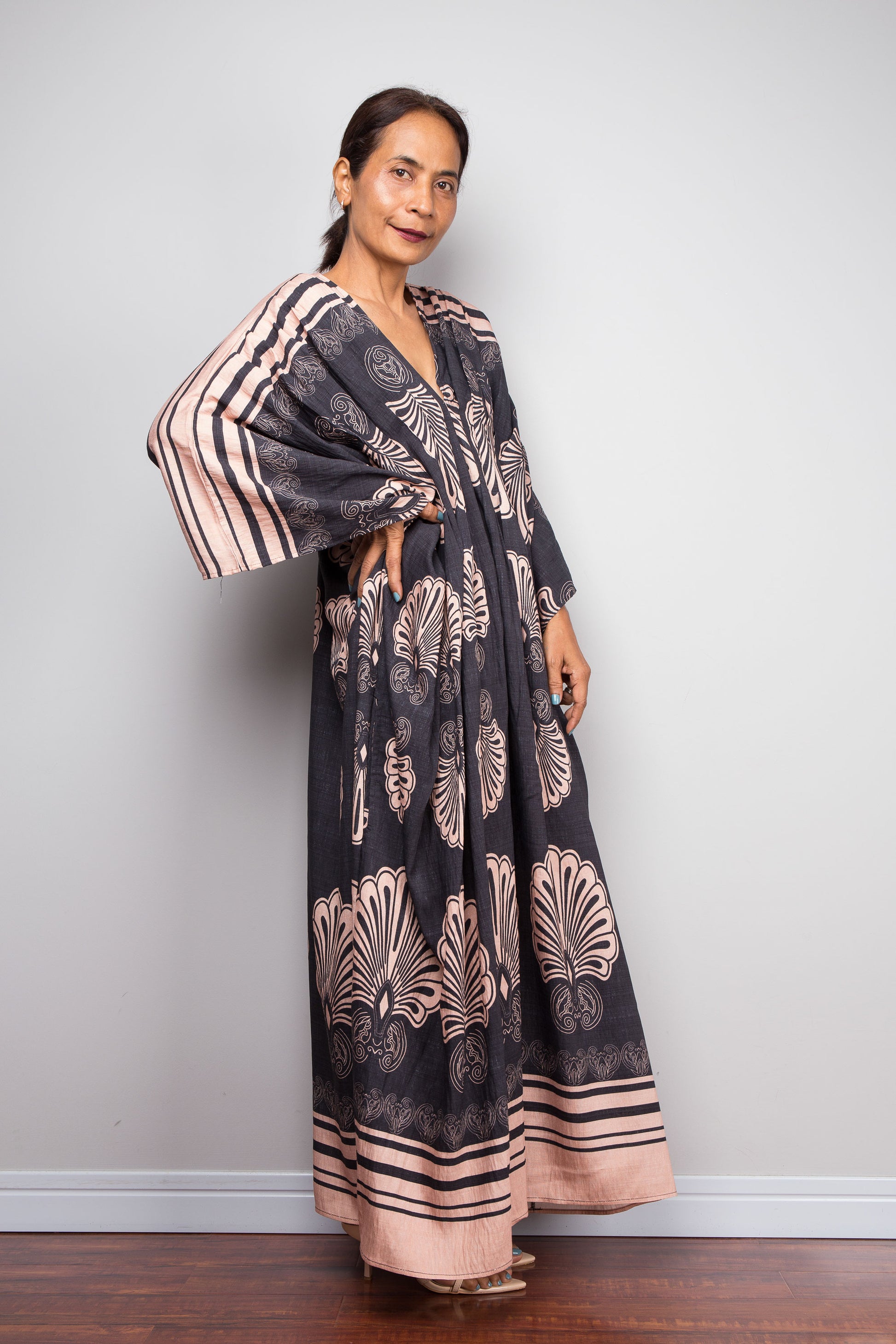 Three quarter Front-facing studio shot of a petite female model in the Nuichan charcoal and blush batwing kaftan dress with hand on hip, displaying the wide kimono sleeves with contrasting underside print, deep V-neckline, and dramatic shoulder pleating