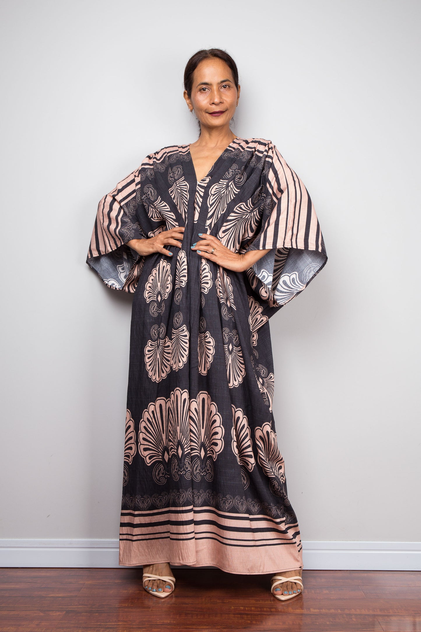 Front-facing studio shot of a model in the Nuichan charcoal and blush batwing kaftan dress with hands on hips, displaying the wide kimono sleeves with contrasting underside print, deep V-neckline, and dramatic shoulder pleating