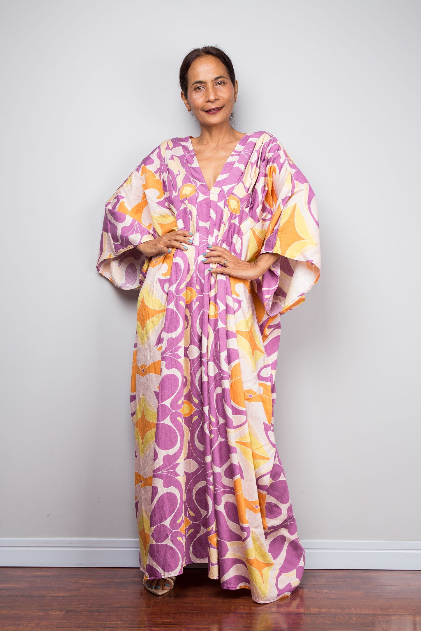 Front facing studio shot of a model in the Nuichan violet and amber batwing kaftan dress with hands on hips, displaying the wide kimono sleeves, deep V neckline, and dramatic shoulder pleating