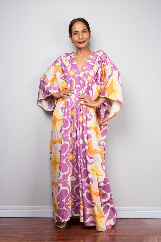 Front facing studio shot of a model in the Nuichan violet and amber batwing kaftan dress with hands on hips, displaying the wide kimono sleeves, deep V neckline, and dramatic shoulder pleating