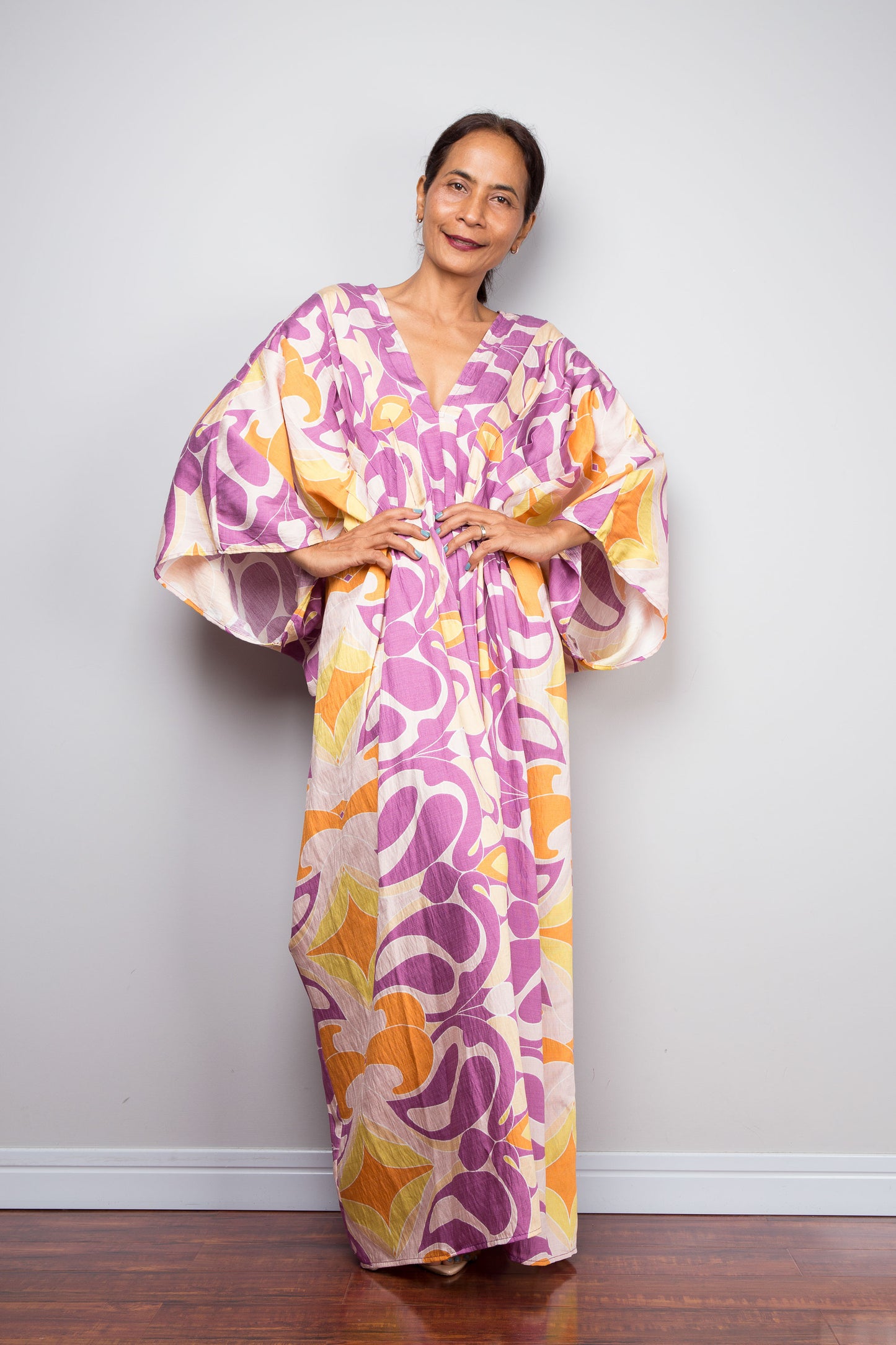 A woman modeling a long Nuichan kaftan dress with a bold abstract floral pattern in purple, orange, and yellow on a cream background, standing against a plain light gray wall.
