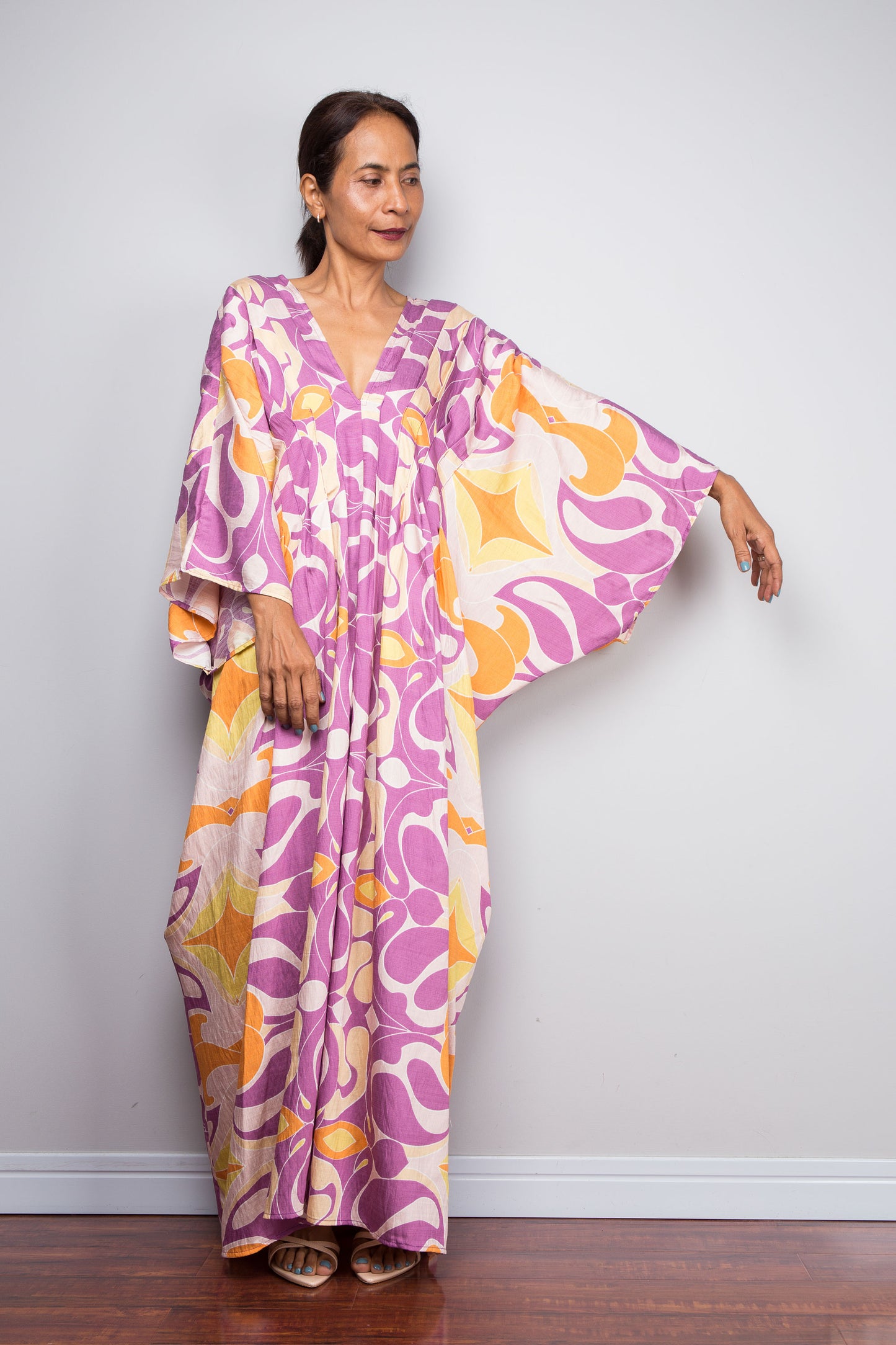 A petite woman modeling a long Nuichan kaftan dress with a retro mid-century abstract pattern in purple, orange, and yellow on a cream background, standing against a light gray wall.