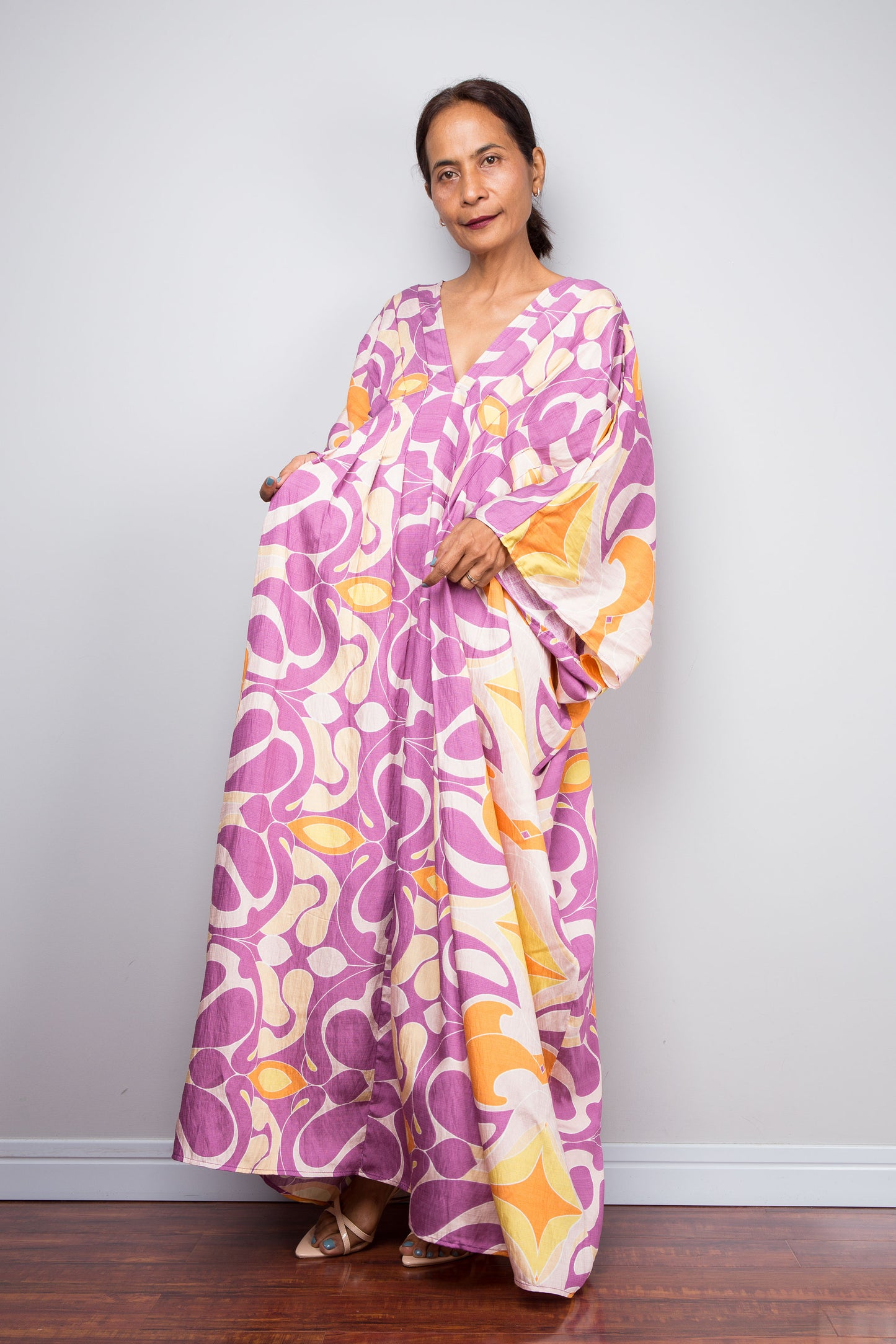 A woman modeling a long, flowing kaftan dress with a retro abstract swirl pattern in purple, pink, orange, and cream, standing against a light gray wall.