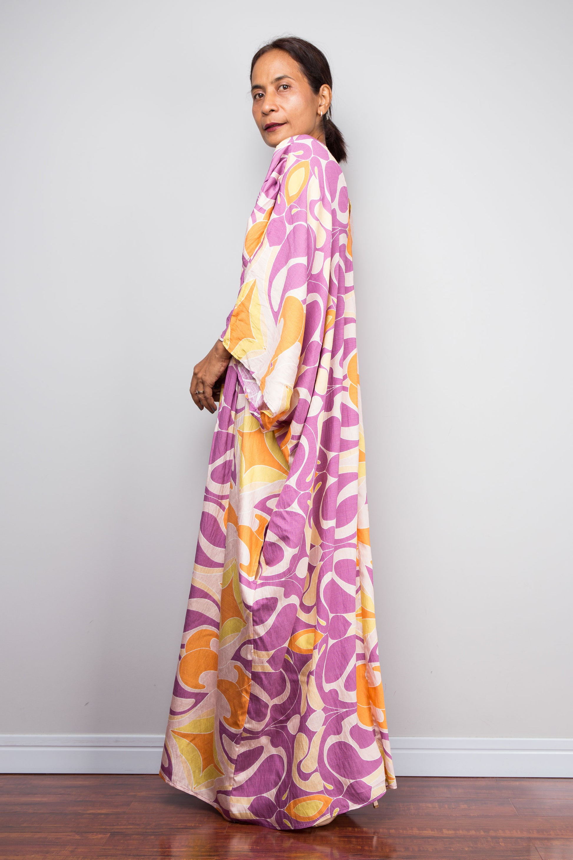 A woman wearing a long, flowing kaftan dress with a bold abstract floral pattern in purple, orange, and cream, standing against a plain light gray wall.