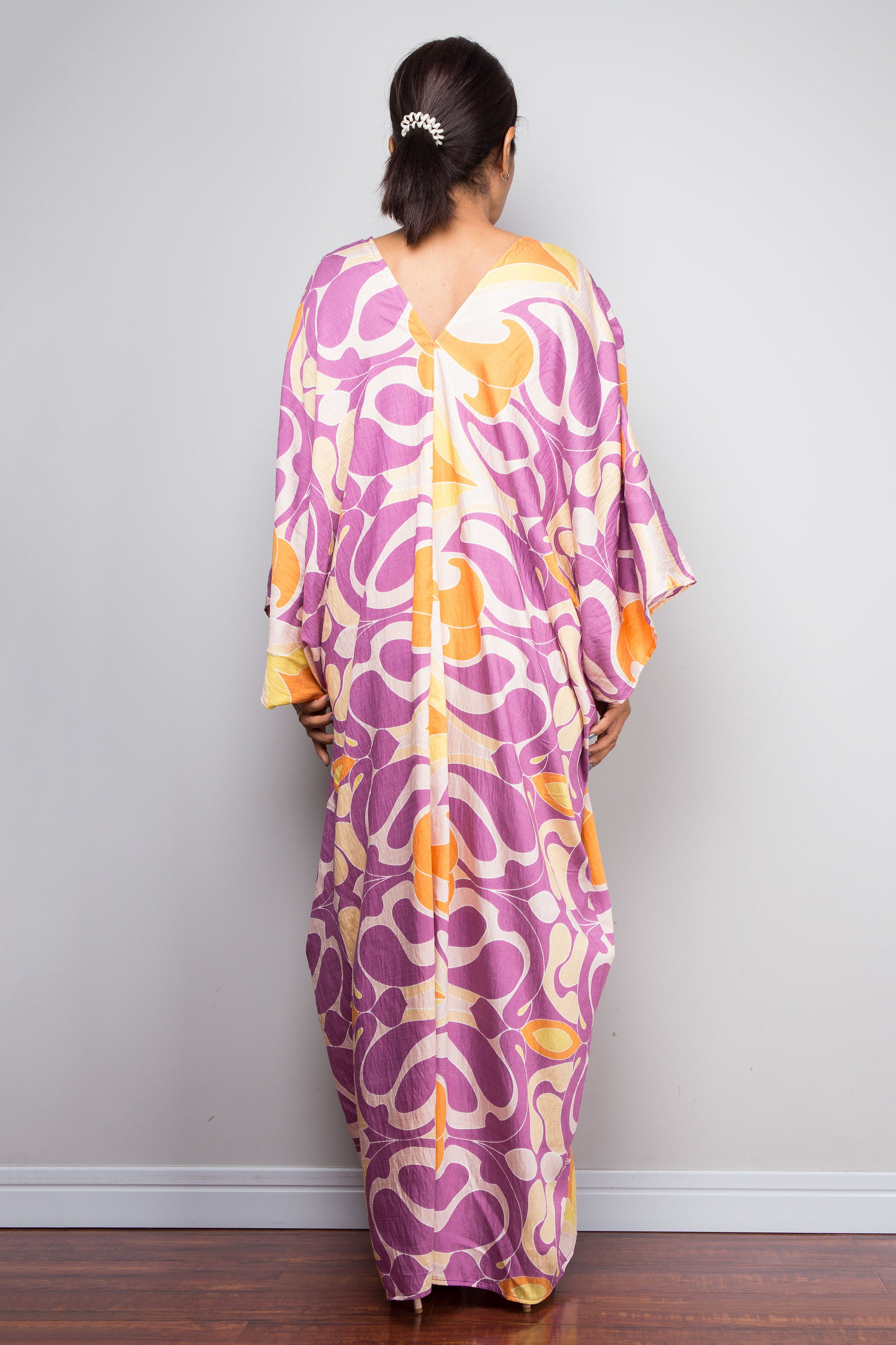 Rear view of a model wearing the Nuichan violet and amber batwing kaftan dress, highlighting the structural shoulder to hem pleating and full length Art Deco swirl print sweeping to the floor