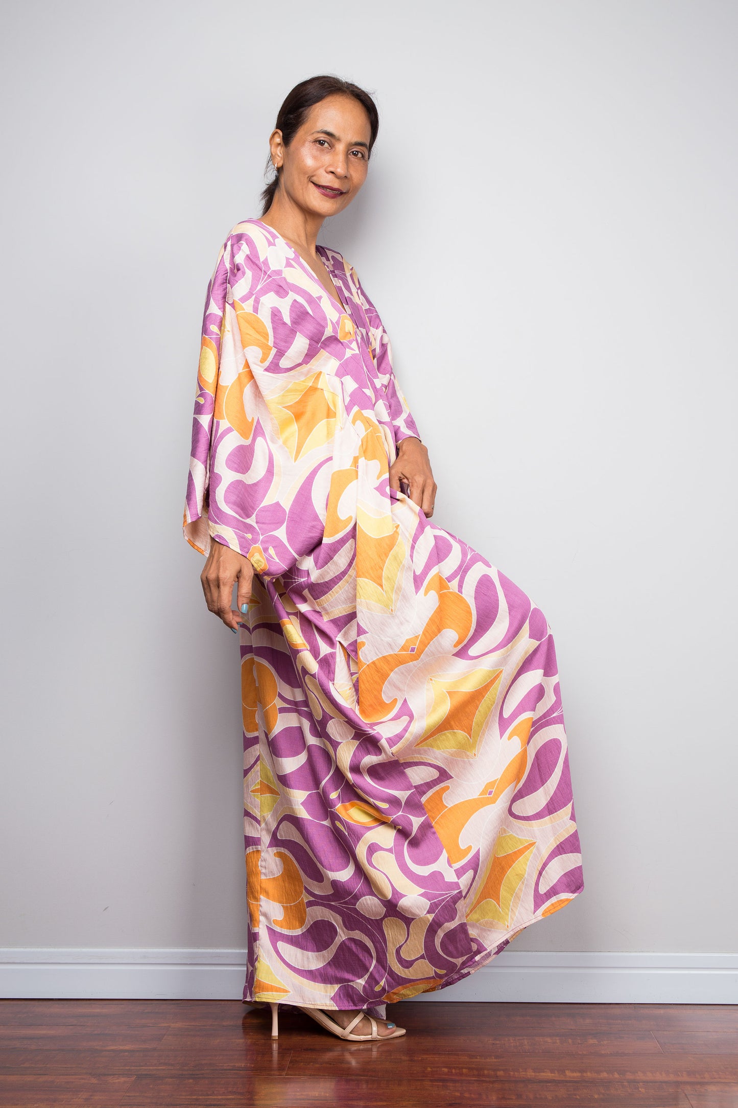 A woman modeling a long, flowing kaftan dress with an abstract wavy pattern in purple, orange, and cream, standing against a light gray wall on a dark wood floor.