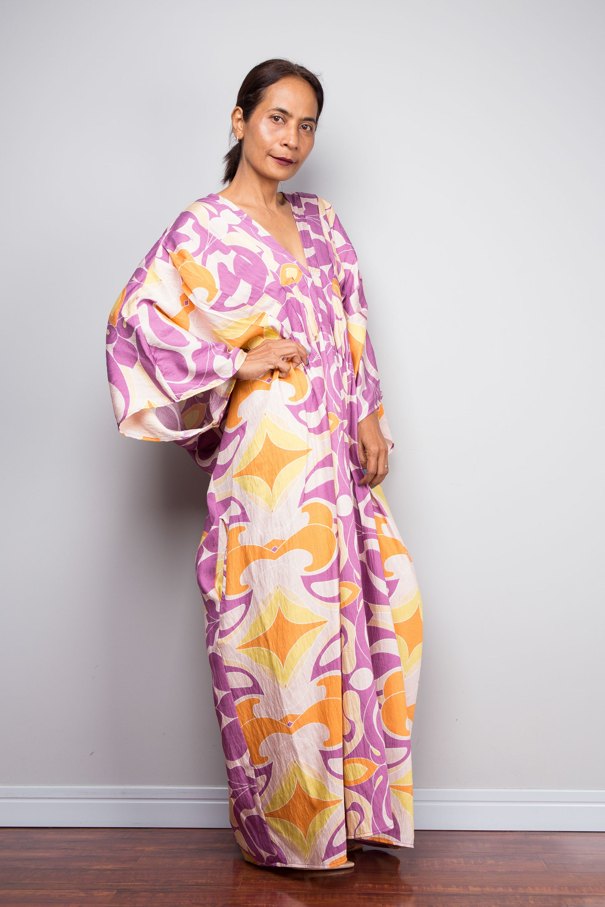 A woman modeling a long, loose kaftan dress with a bold abstract floral pattern in purple, orange, and cream, standing against a plain light gray wall.