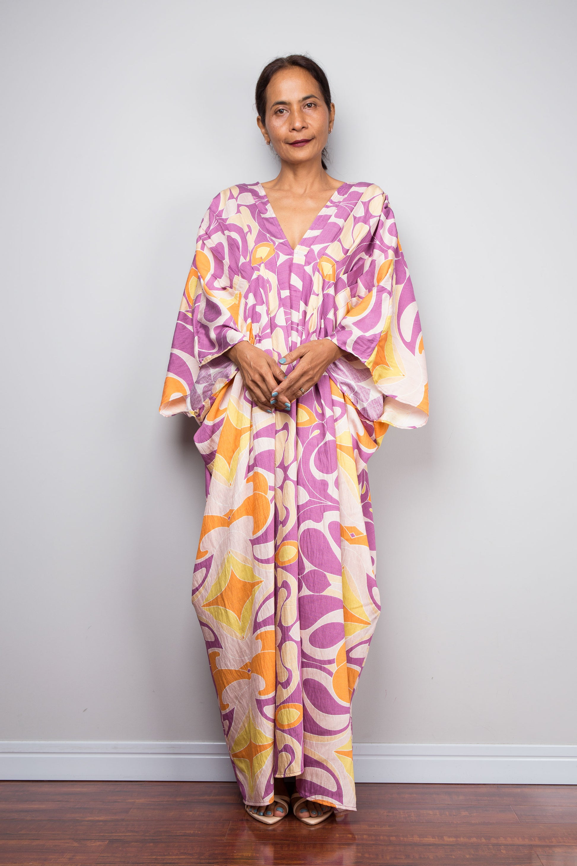 A woman wearing a loose, long kaftan dress with a vibrant purple, orange, and cream abstract floral pattern, standing against a plain light gray wall.