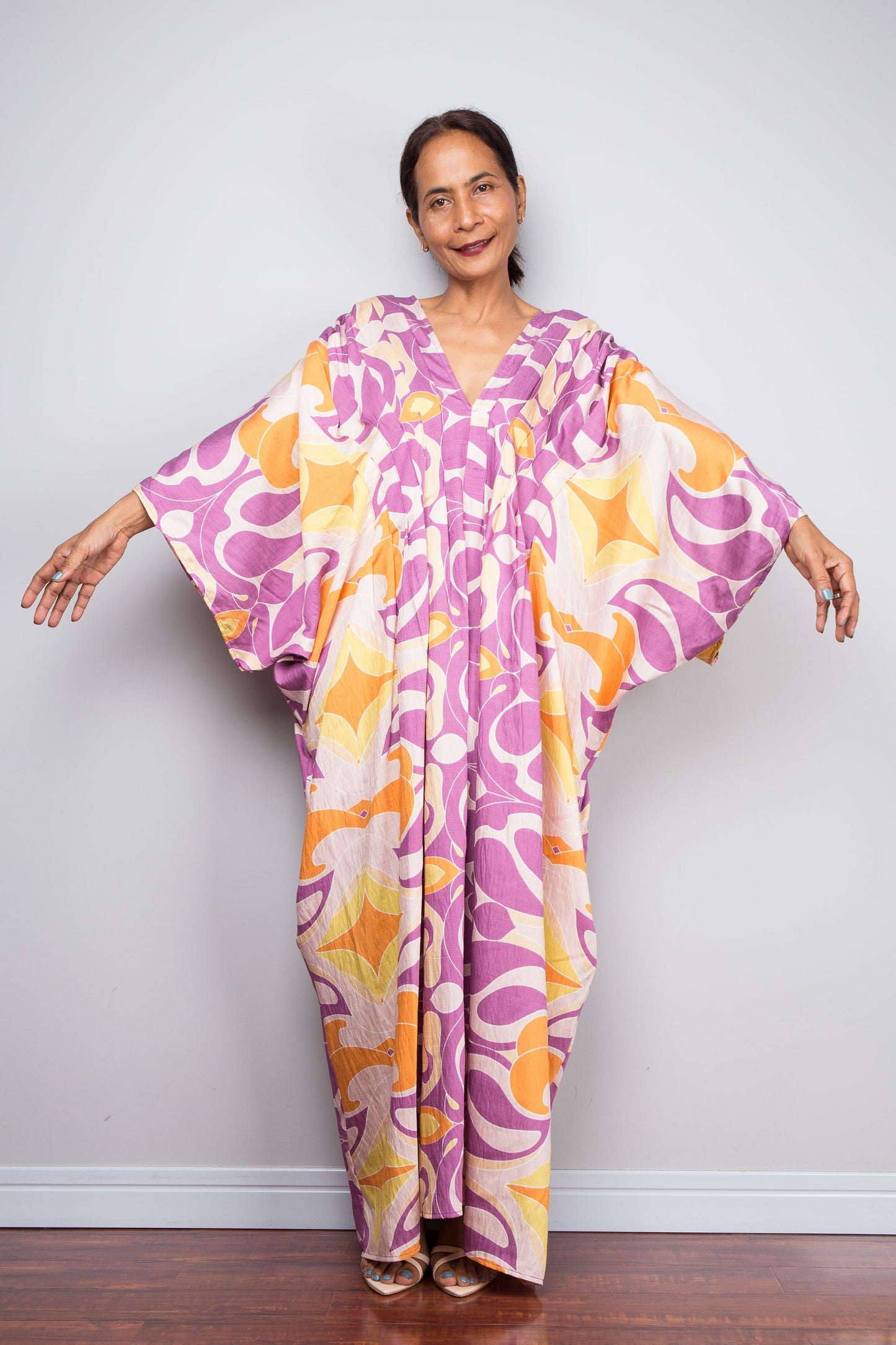 Front facing studio shot of a model in the Nuichan violet and amber batwing kaftan dress with arms extended, showcasing the full sweep of the wide kimono sleeves and bold swirl print