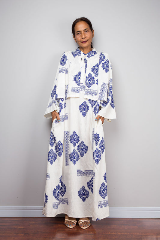 Front-facing view of a model wearing the Nuichan white and blue medallion print kaftan maxi dress with attached capelet and hands in side pockets, showcasing the cobalt blue diamond medallion damask print, horizontal border banding, capelet layer over the upper body, mandarin collar with three-button placket, and long sleeves with flared cuffs