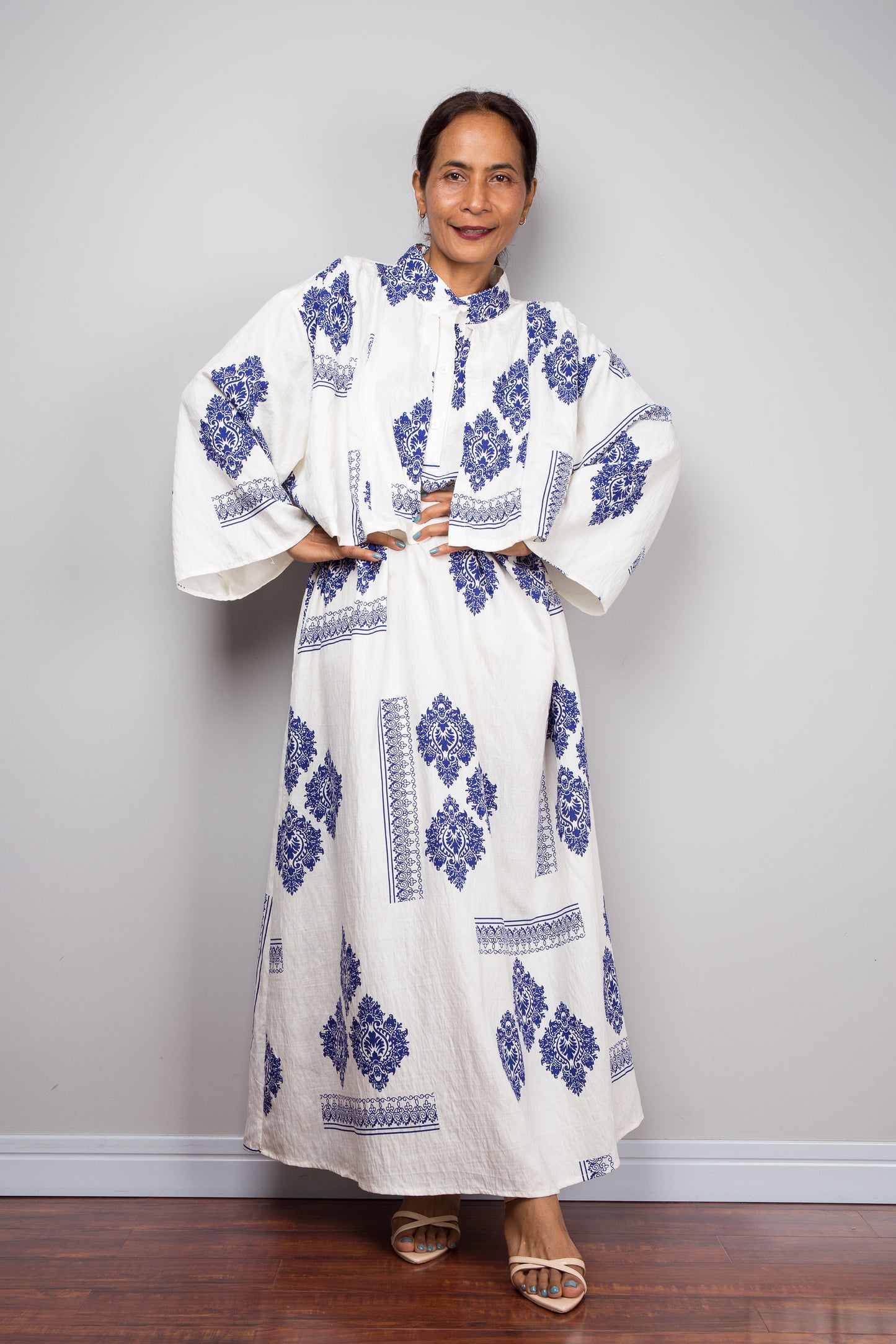 A woman modeling a white caftan maxi dress with blue floral and geometric patterns, standing against a light gray wall on a wooden floor.